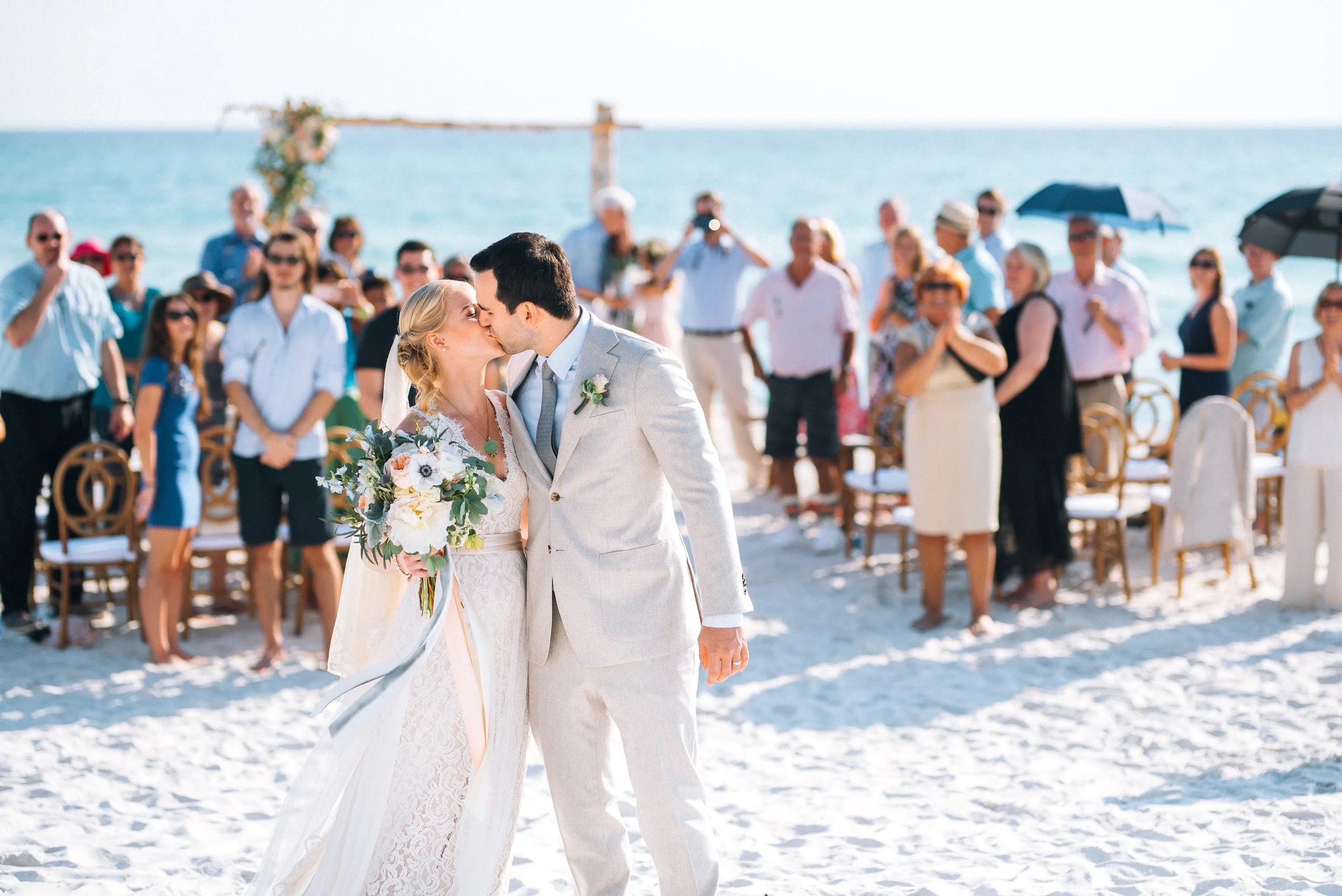 A couple is kissing during a wedding ceremony on a beach, surrounded by guests and ocean on their wedding day in Florida 