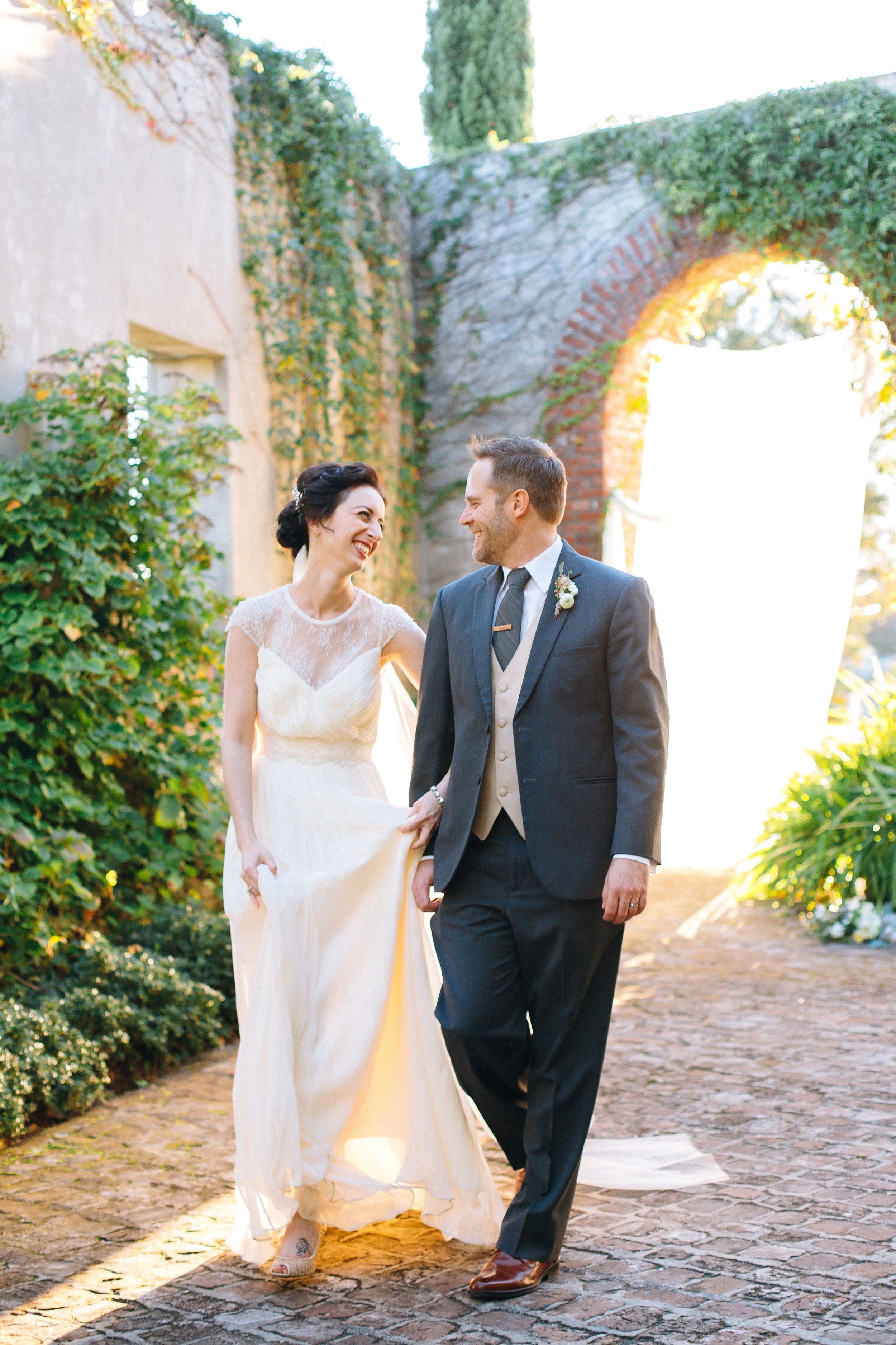 A bride and groom walking outdoors, smiling and holding hands, with a rustic brick and stone background covered in ivy and greenery at Summerhill Studios in Atlanta Wedding photography by Sowing Clover Photography