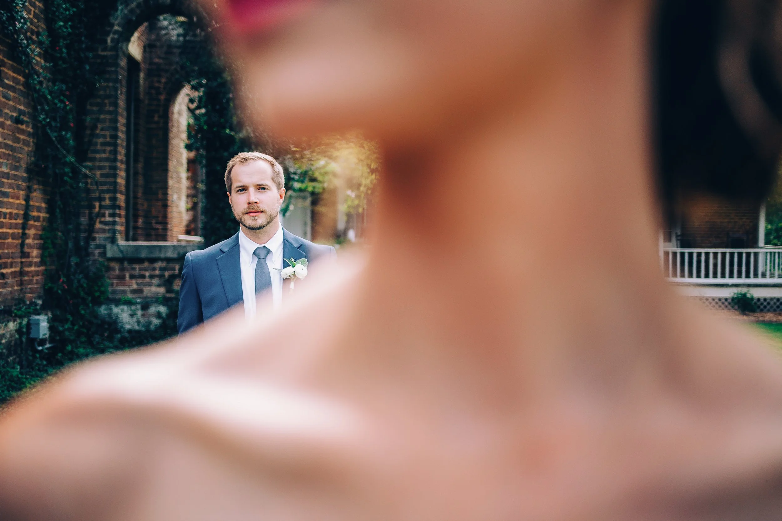 A groom in a suit and tie stands outdoors near a brick building, looking directly at the camera, shot through the chin and shoulder of a bride in foreground.  Shot at Barnsley Resort in North Georgia. Wedding photography by Sowing Clover Photography.