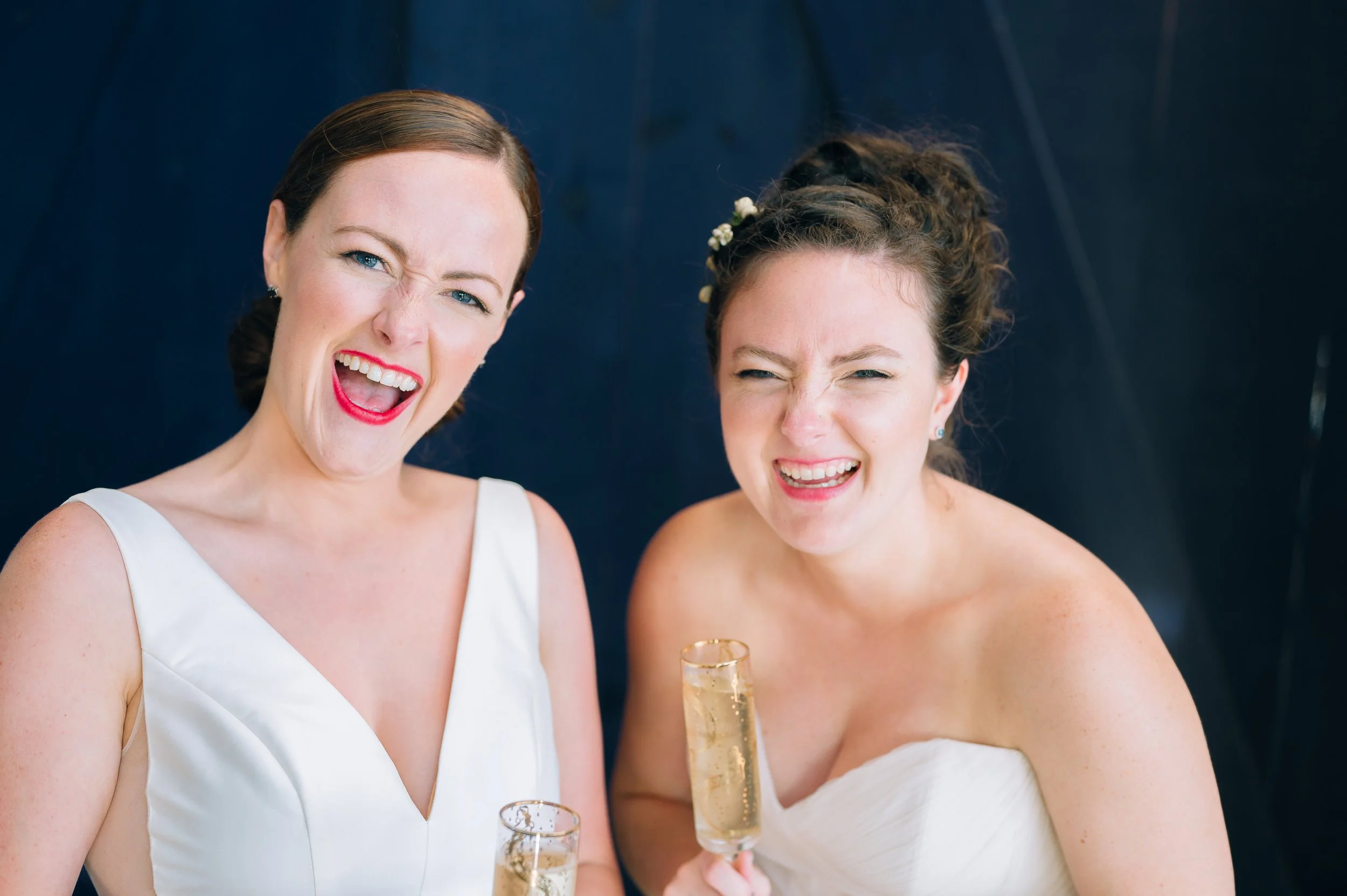 Two women wearing white dresses, celebrating with glasses of champagne and making cheerful faces, against a dark background. Wedding Photography by Sowing Clover Photography.