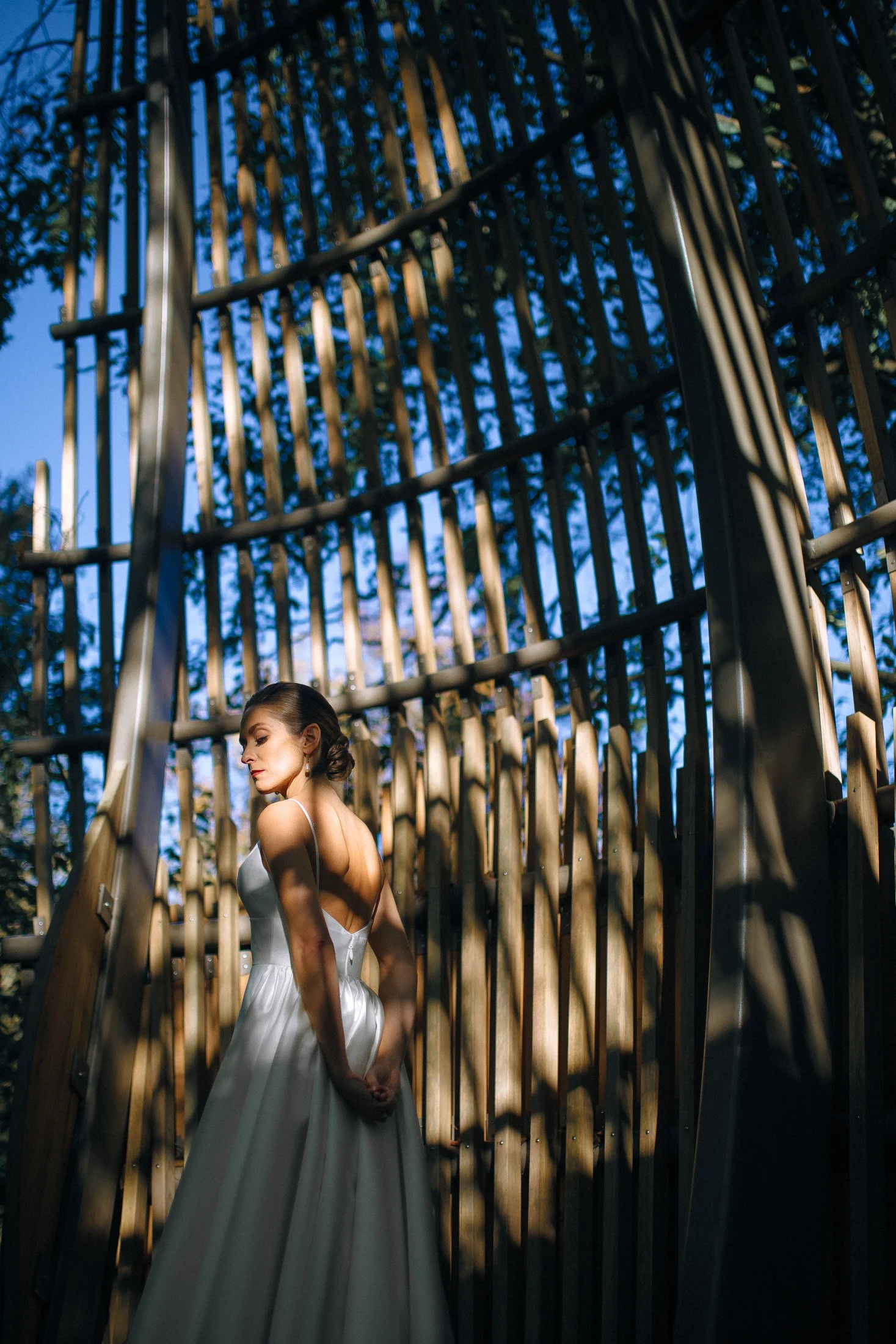 A bride in a white dress standing near a wooden fence with shadows cast by trees, during daytime at the Fernbank Science Center in Atlanta. Wedding Photography by Sowing Clover Photography.