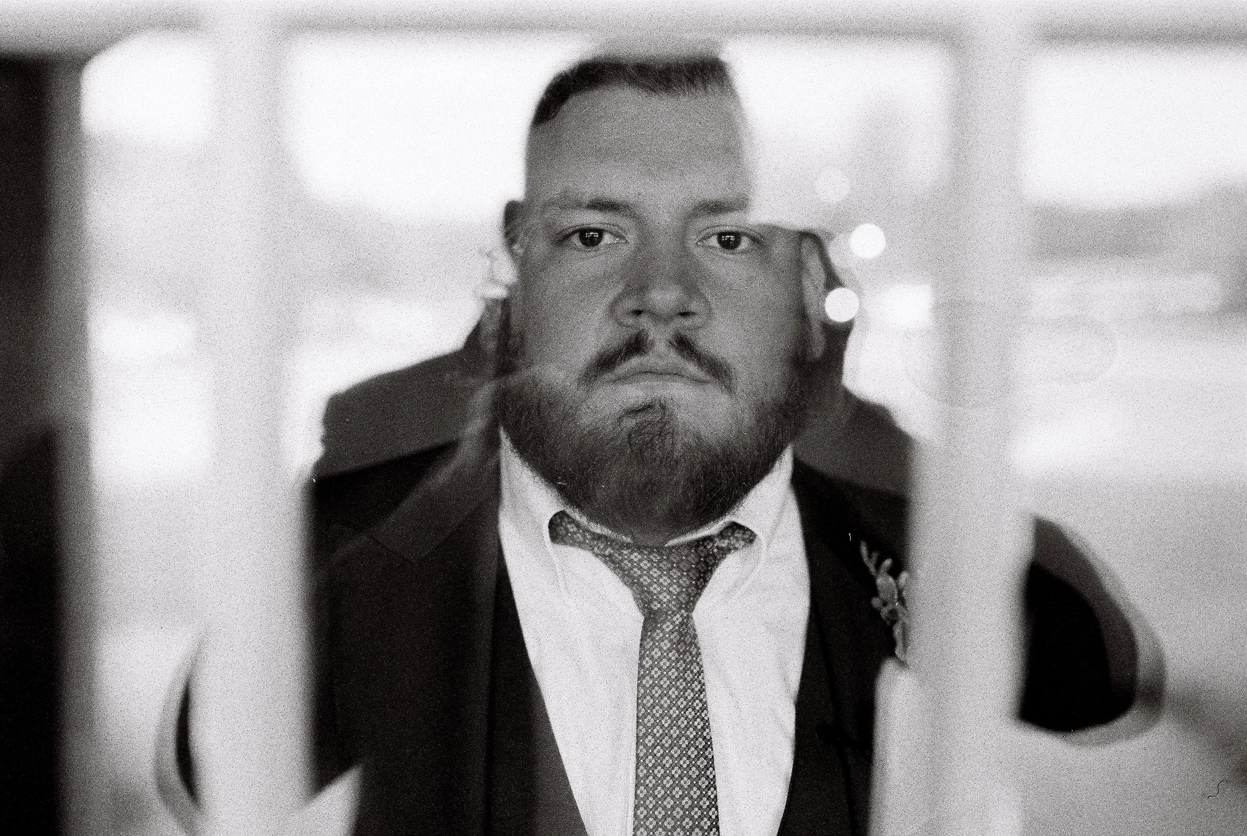 A black and white photo of a man with a beard, wearing a suit and tie, looking directly at the camera through what appears to be a window or glass barrier at Mint Springs Farm, Nashville. Wedding Photography in by Sowing Clover Photography