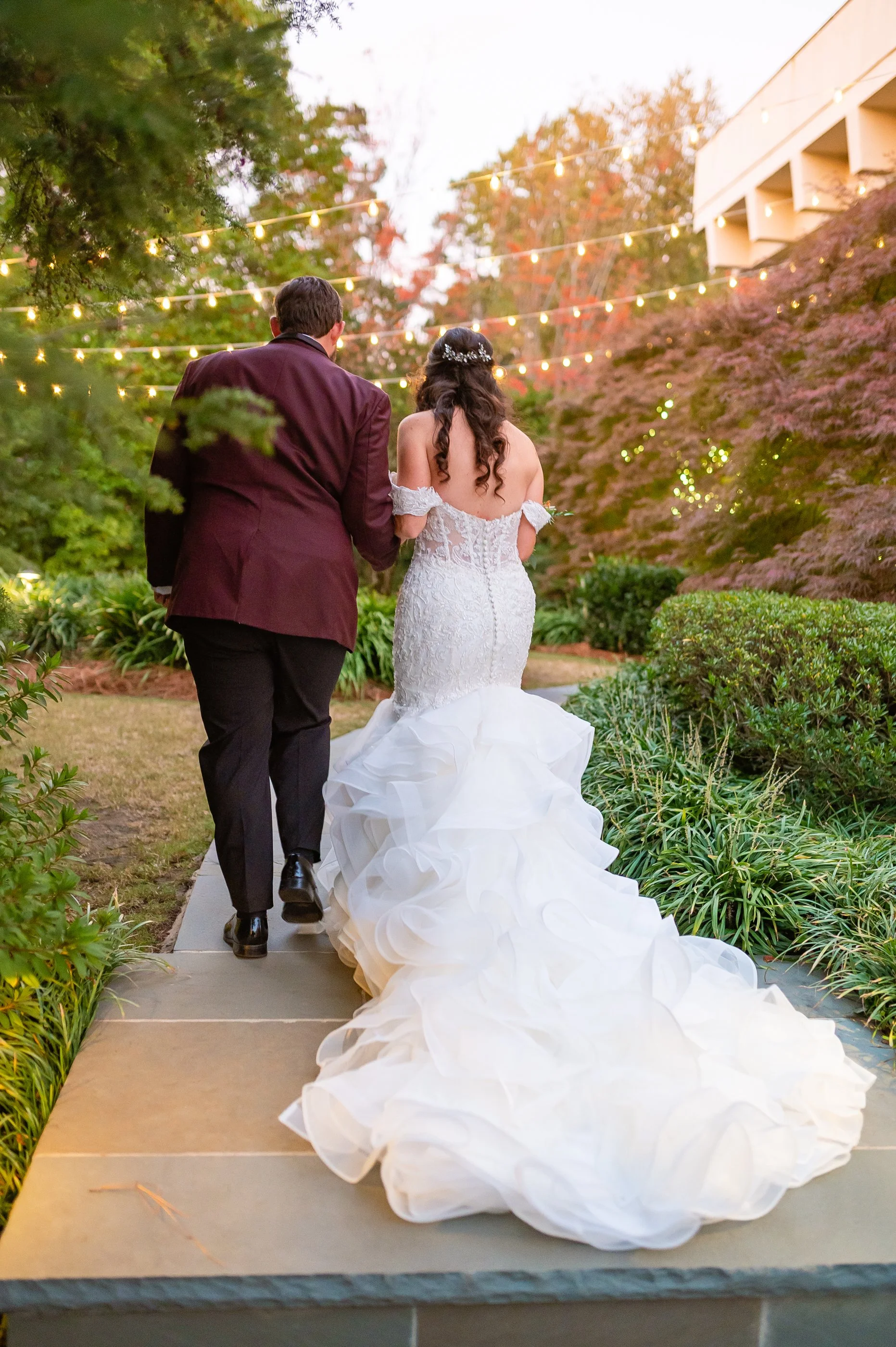 A bride and groom walking hand in hand on a garden pathway during their wedding celebration at dusk, with string lights overhead and colorful trees in the background at The Atrium in Norcross GA. Wedding Photography by Sowing Clover Photography.