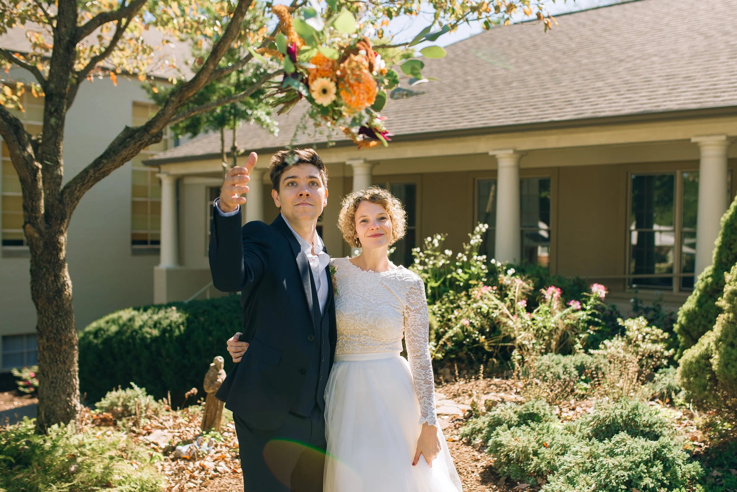 A newlywed couple is throwing a bouquet in the air outdoors in front of a house with garden and trees on a sunny day. Wedding Photography in Nashville by Sowing Clover Photography