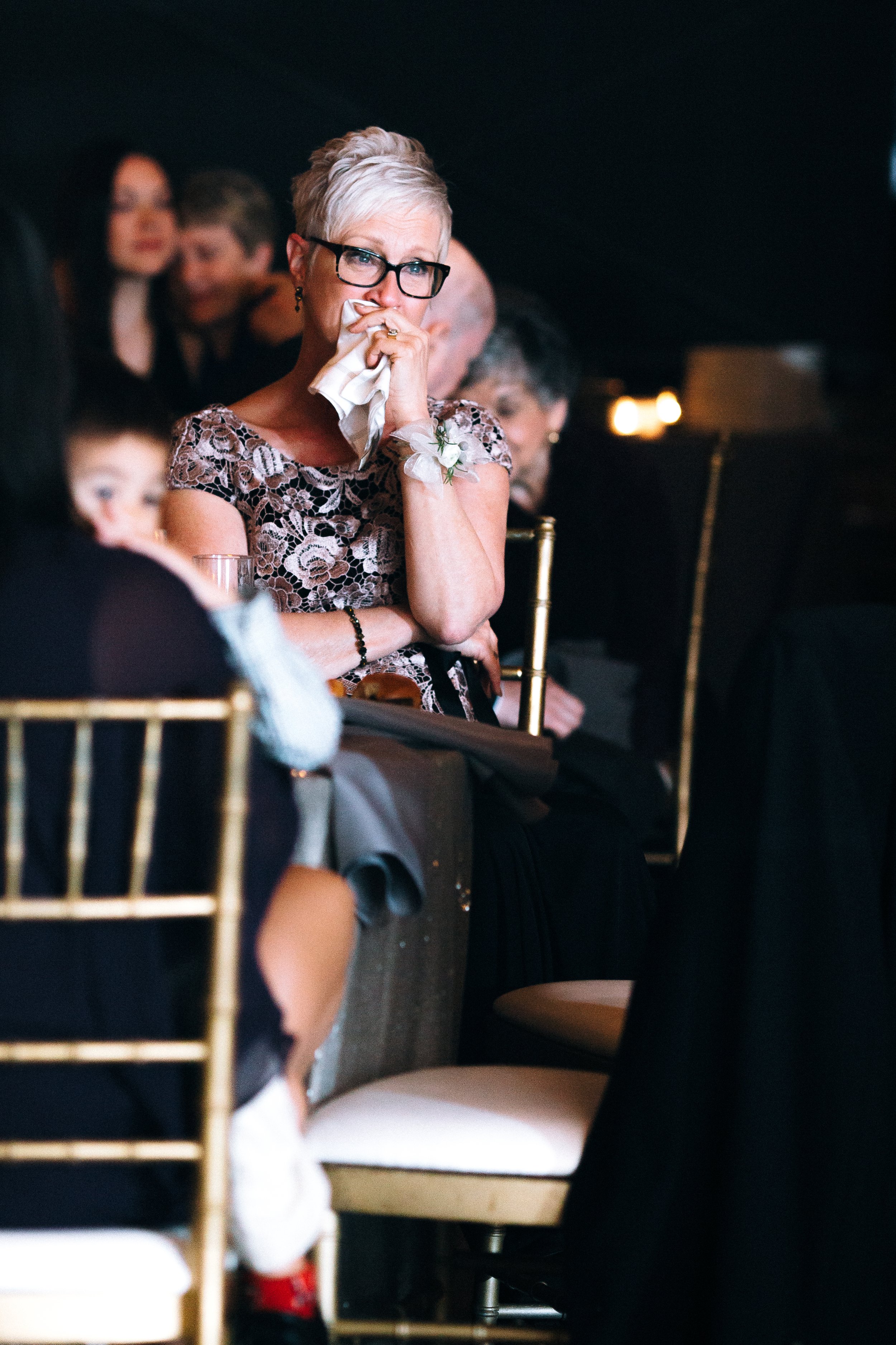 A woman with short gray hair, glasses, and a floral lace dress is sitting at an event, holding a tissue to her face, appearing emotional. at Summerhill Studios in Atlanta Wedding photography by Sowing Clover Photography.