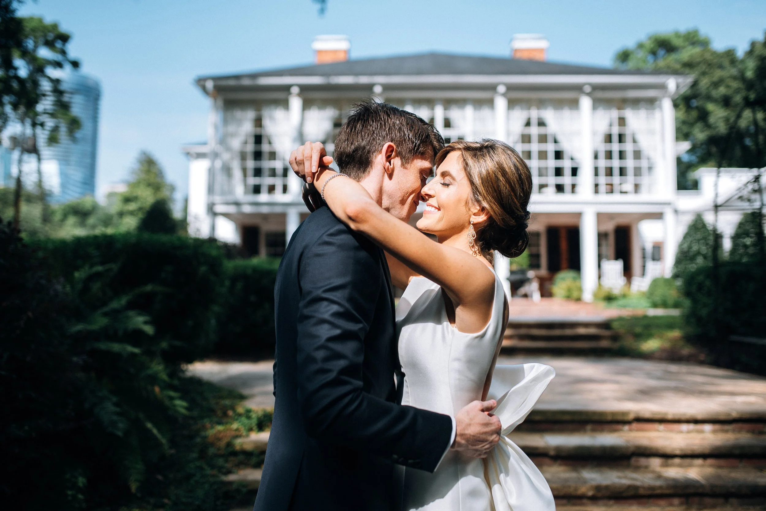 A bride and groom embrace and smile in front of a large white house with steps and surrounded by greenery at The Estate on Piedmont Road in Atlanta, GA.  Wedding Photography by Sowing Clover Photography.