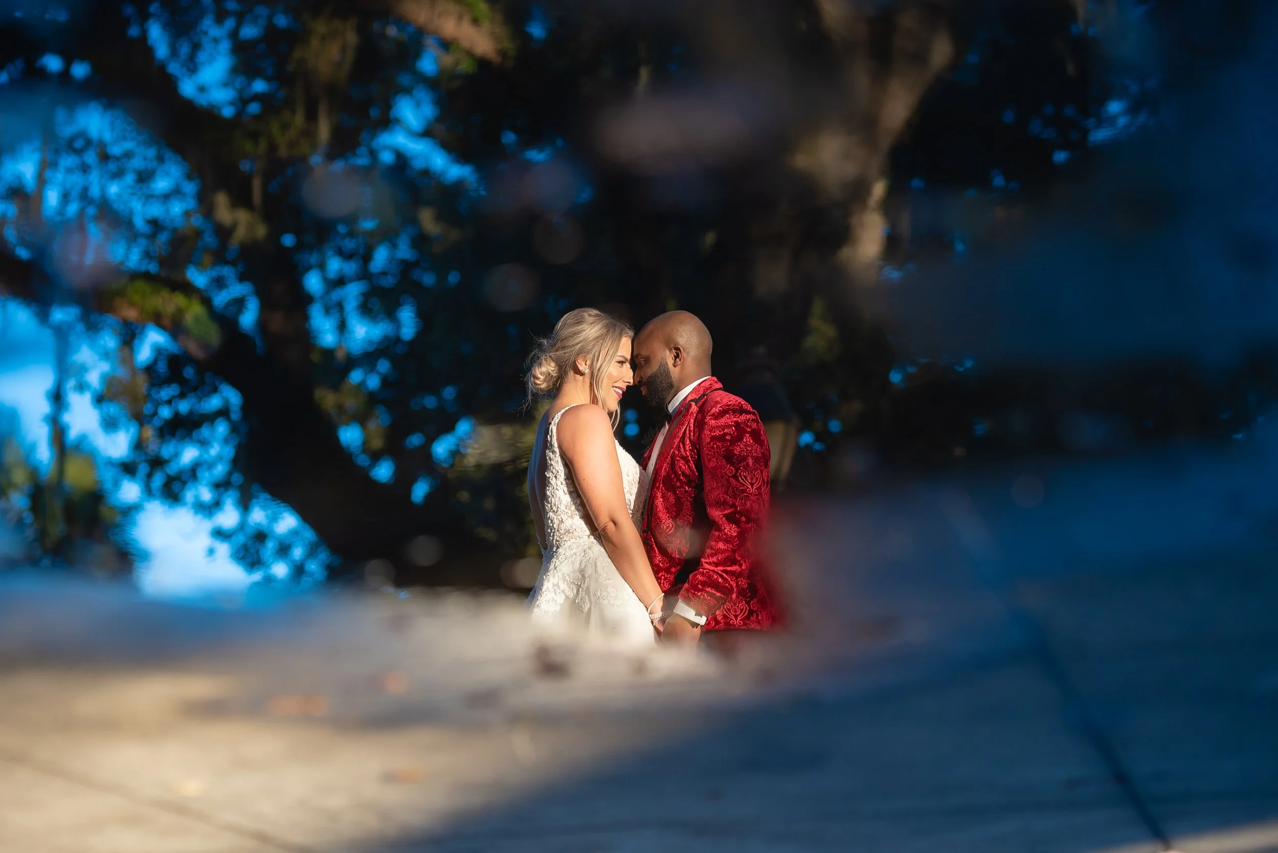 A bride and groom standing close together, touching foreheads and smiling, outdoors with a large tree and blue sky in the background during sunset at Forsyth Park in Savannah, Georgia. Wedding Photography by Sowing Clover Photography.