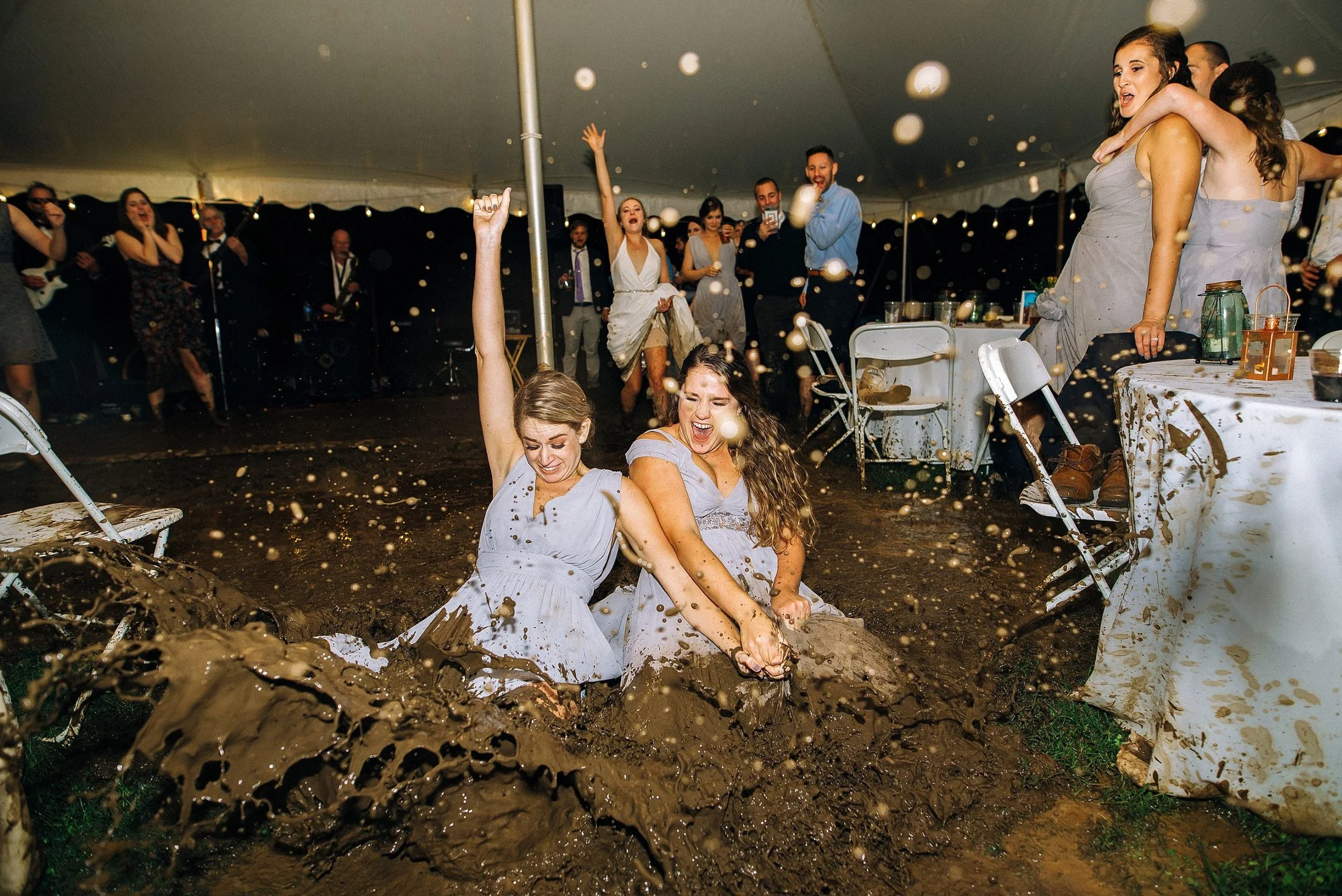 Two bridesmaids in light-colored dresses are seated in mud under a large tent at a celebration, splashing mud with joyful expressions, while guests around them cheer and enjoy the event. Nashville. Wedding photography by Sowing Clover Photography