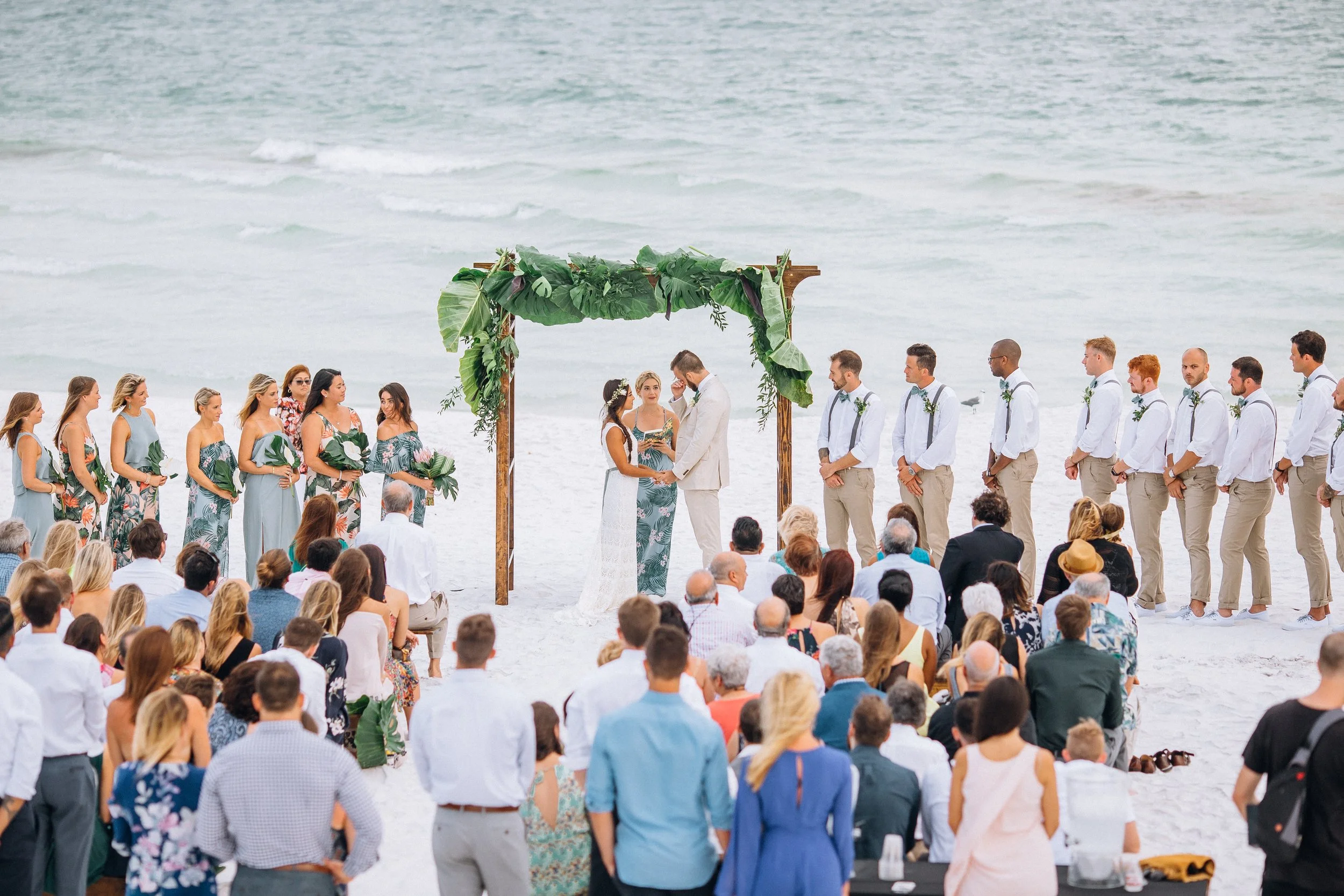 Beach wedding ceremony with couple exchanging vows under a greenery arch, surrounded by bridesmaids and groomsmen, with guests seated on the sand and ocean in the background in Florida. Wedding Photography in by Sowing Clover Photography.