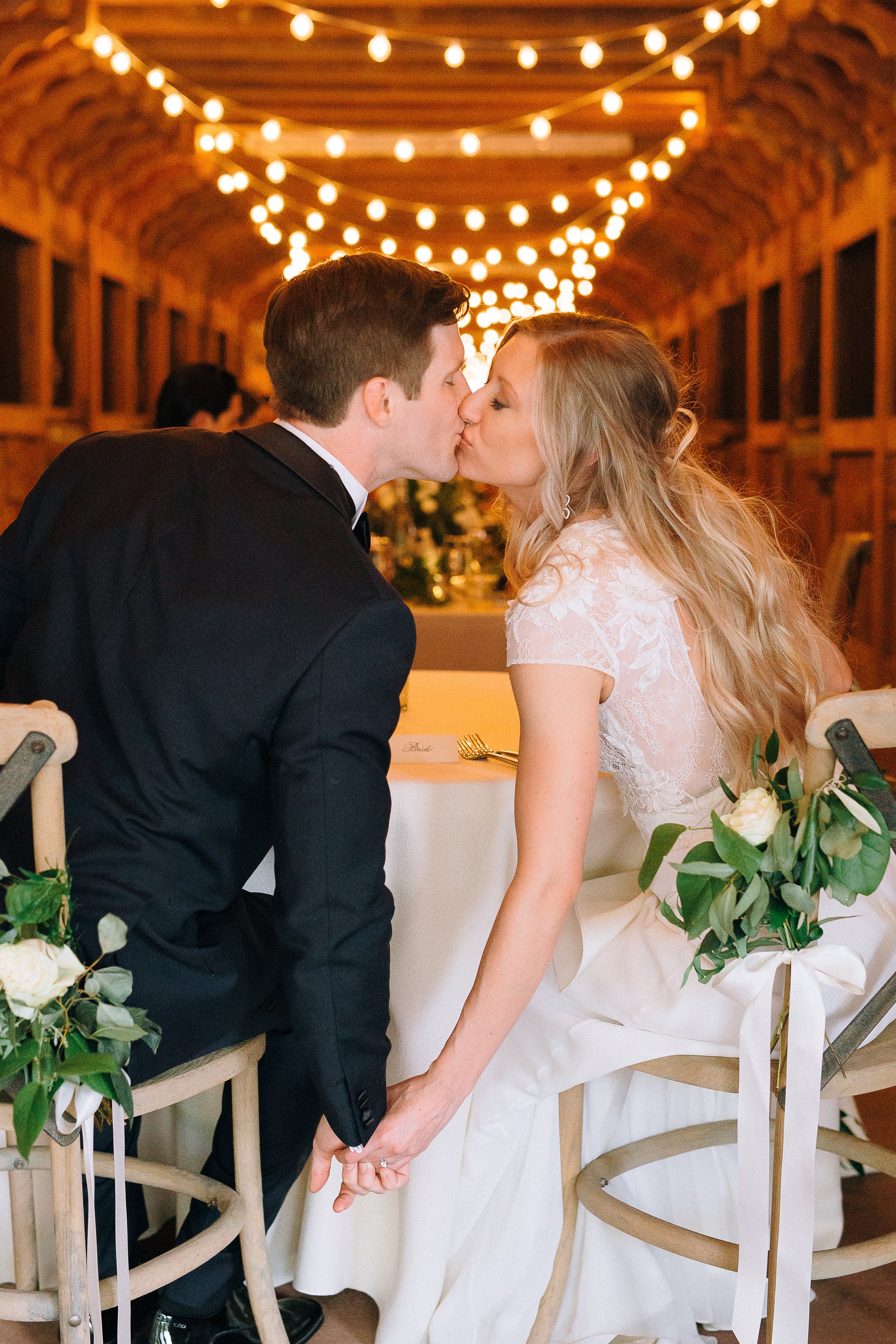 A bride and groom sharing a kiss at their wedding reception, sitting on chairs holding hands, decorated with greenery and flowers, under string lights in a venue at The Farm in Rockmart Georgia Wedding Photography in by Sowing Clover Photography