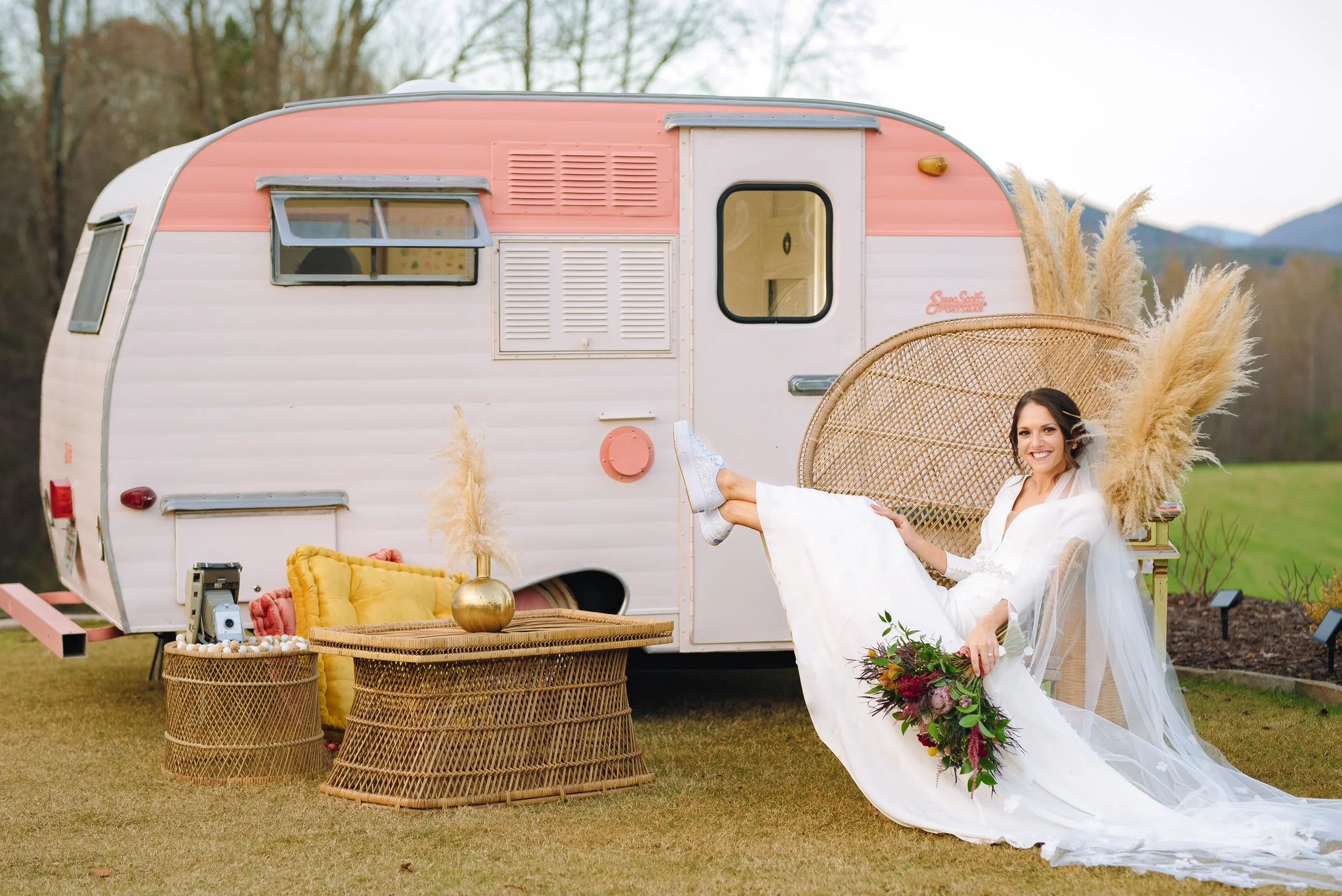 A bride in a white wedding dress sitting on a wicker chair outside near a vintage camping trailer, holding a bouquet of flowers, with decorative vases and pillows around her. North Georgia Wedding.  Wedding Photography by Sowing Clover Photography.
