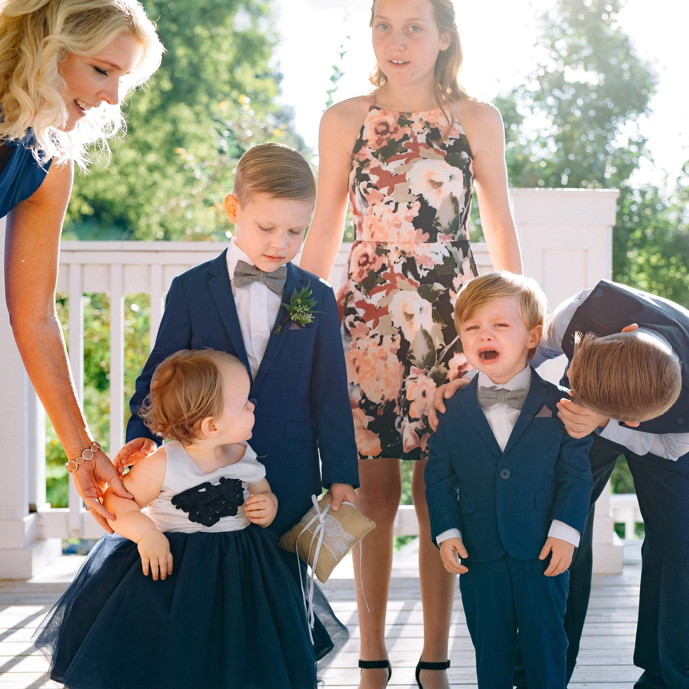 A woman and five young children, dressed in formal attire, are outdoors on a wooden deck with a white railing. The woman is smiling, and one of the children is crying. Mint Springs Farm Nashville.  Wedding photography by Sowing Clover Photography.