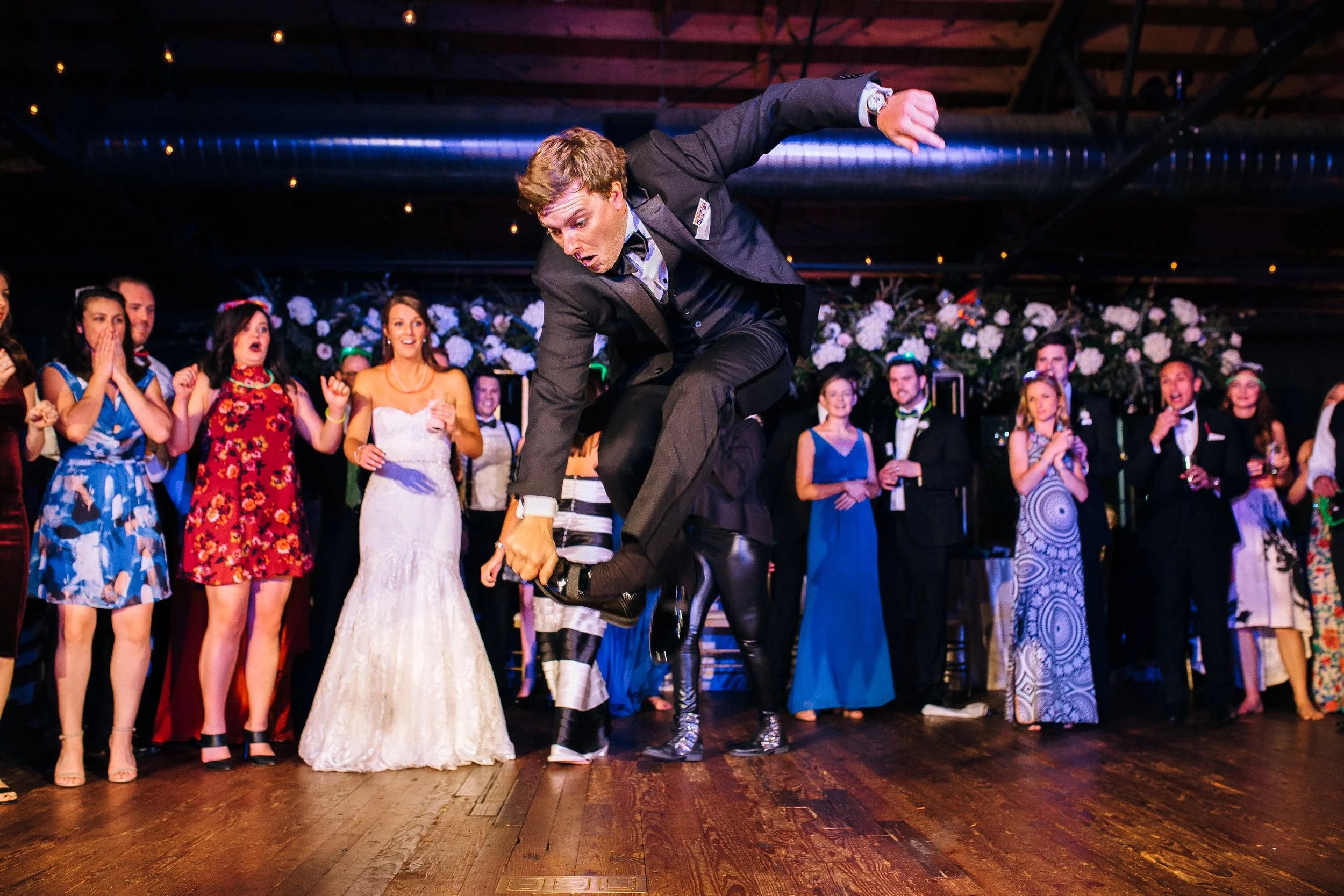 A man in a tuxedo jumping on a dance floor during a celebration, with a group of people watching and reacting joyfully in the background at Summerhill Studios in Atlanta. Wedding Photography in by Sowing Clover Photography