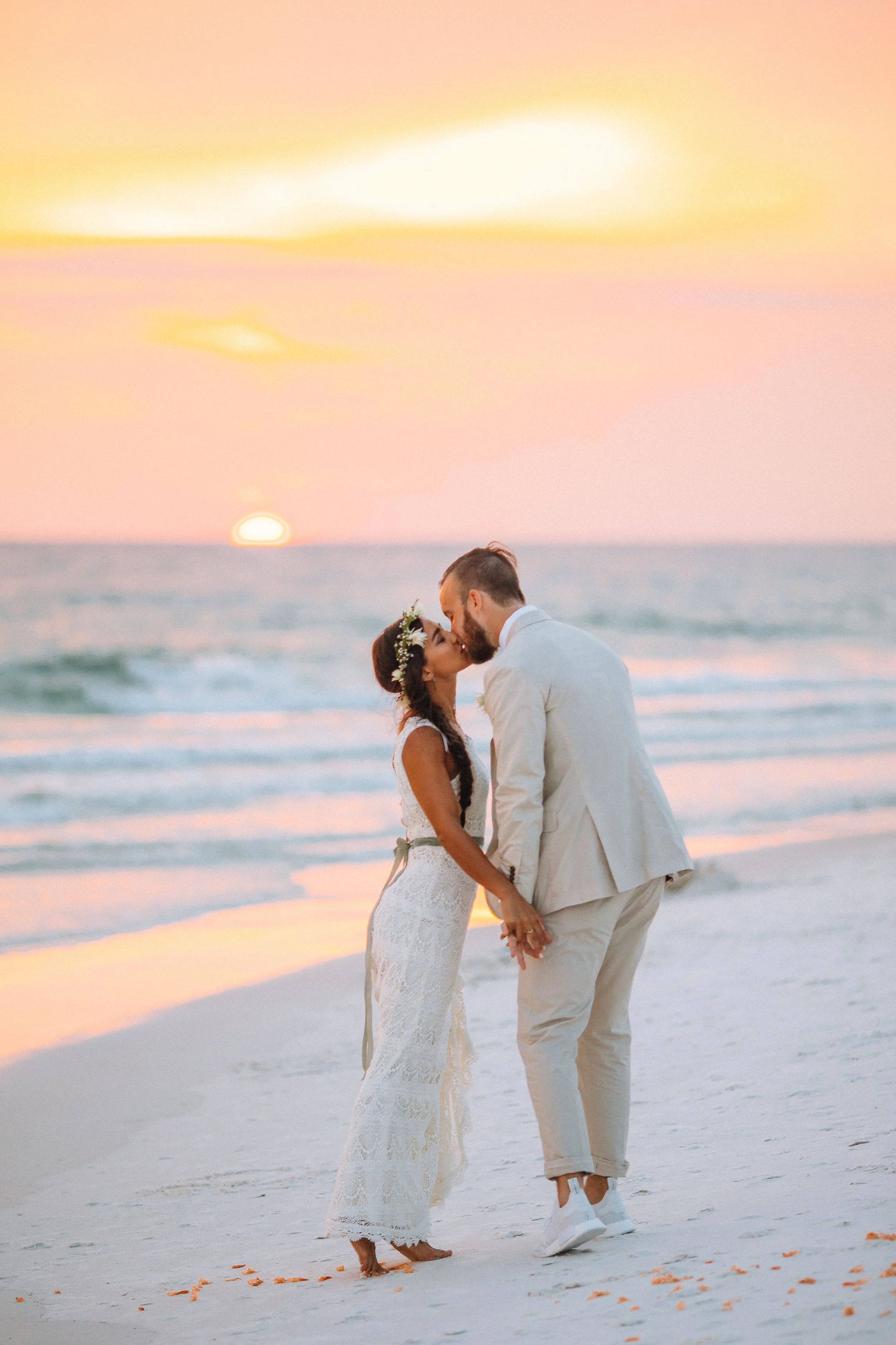 A couple dressed in wedding attire kissing on the beach at sunset, with the ocean and colorful sky in the background in Florida for a beach wedding. Wedding Photography in by Sowing Clover Photography