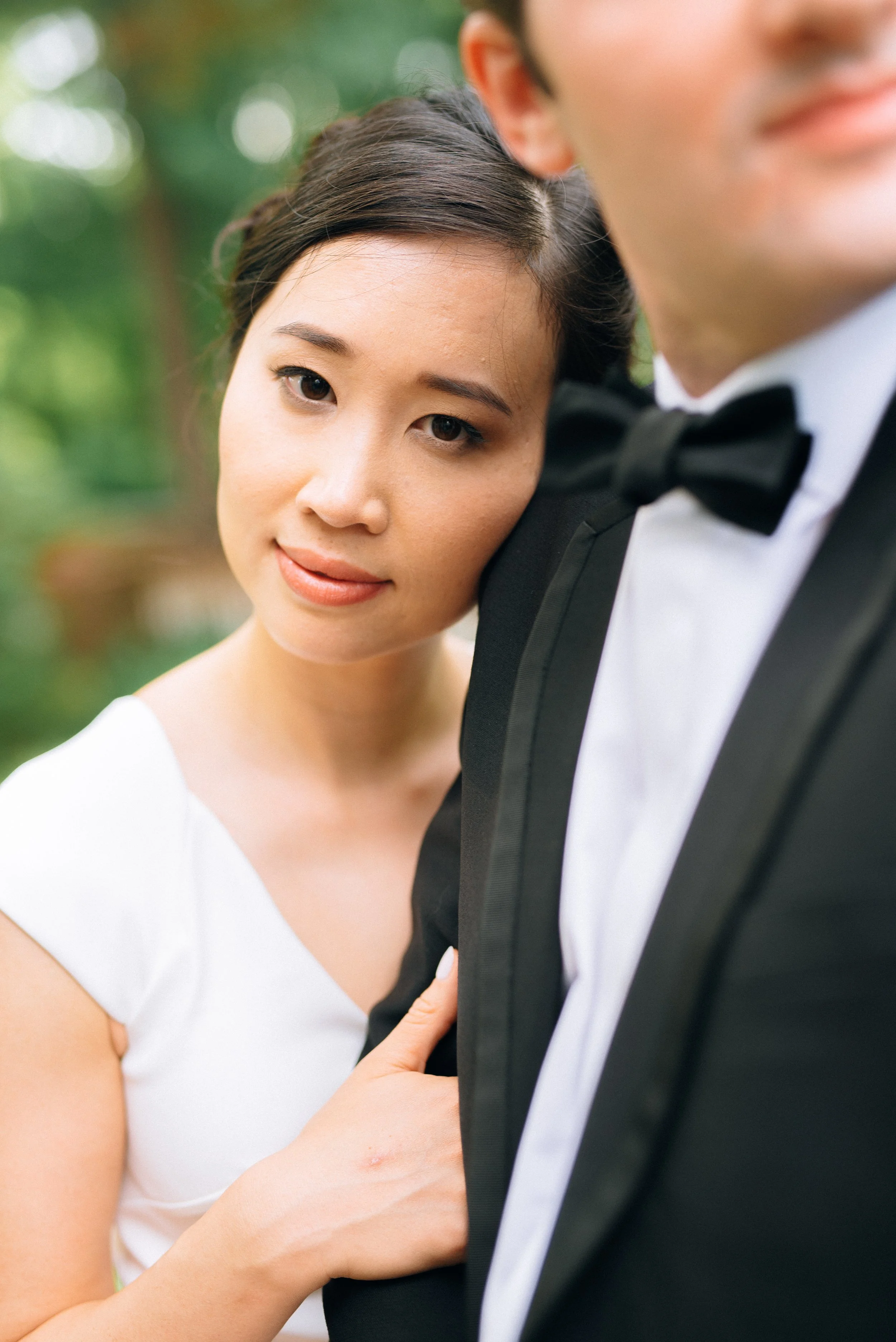 A bride with dark hair in an updo, wearing a white wedding dress, rests her head on a groom in a black tuxedo with a bow tie, outdoors with blurred green foliage in the background in Atlanta. Wedding Photography by Sowing Clover Photography.