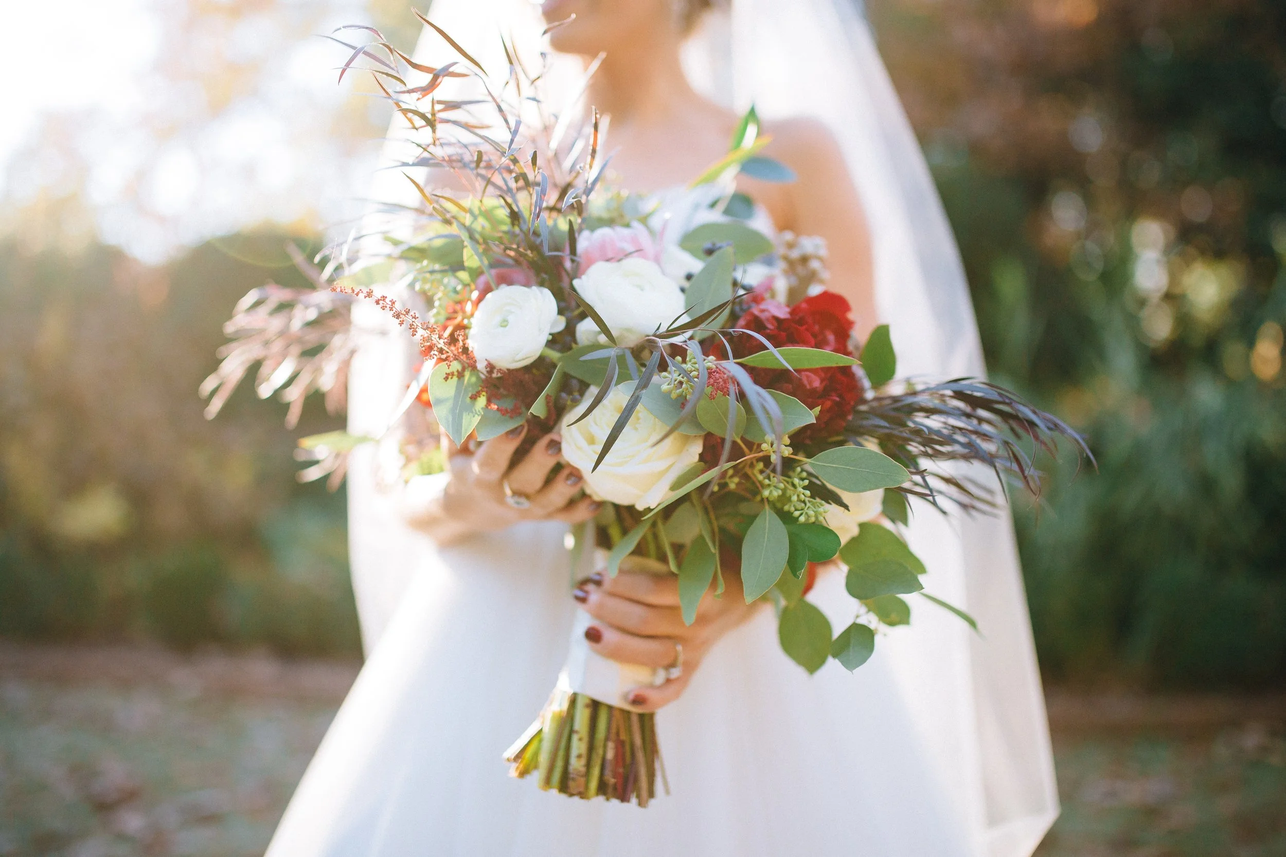 A bride holding a bouquet of white, red, and pink flowers with greenery, outdoors with blurred trees in the background in Atlanta. Wedding Photography by Sowing Clover Photography.
