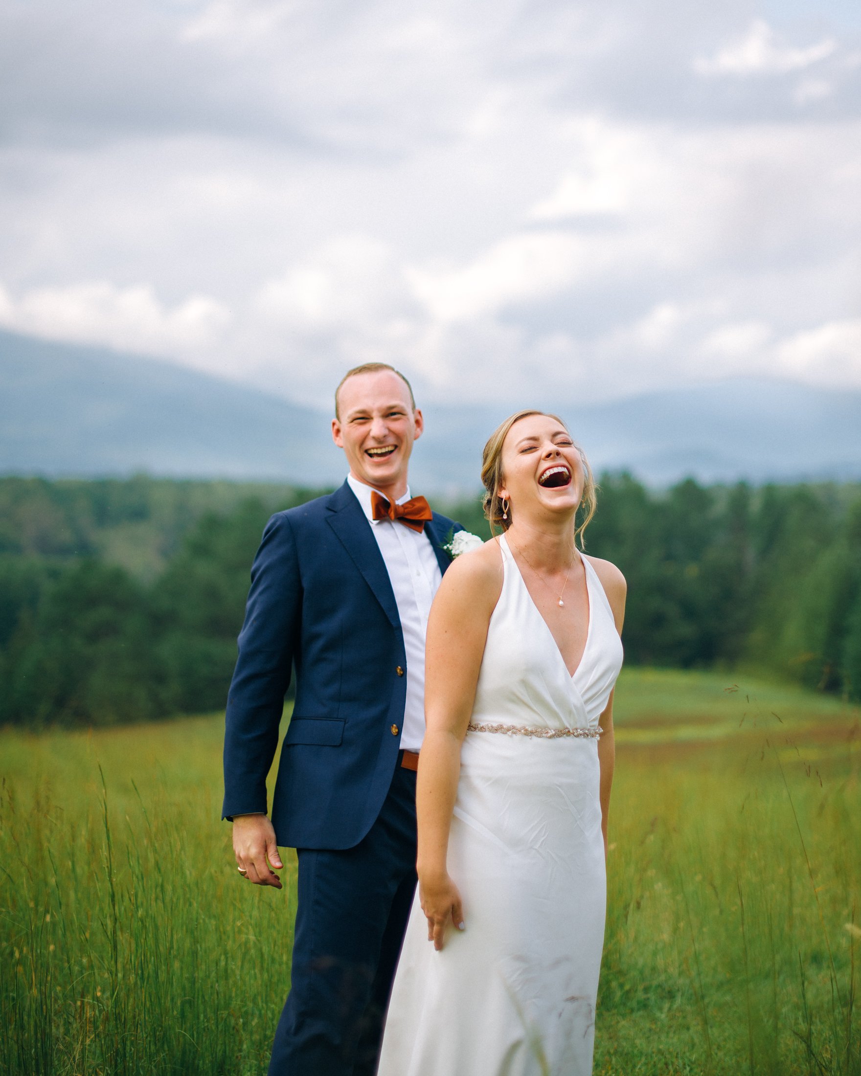 A smiling bride and groom standing in a grassy field outdoors, with a background of trees and mountains under a cloudy sky. In Virginia. Wedding Photography by Sowing Clover Photography.