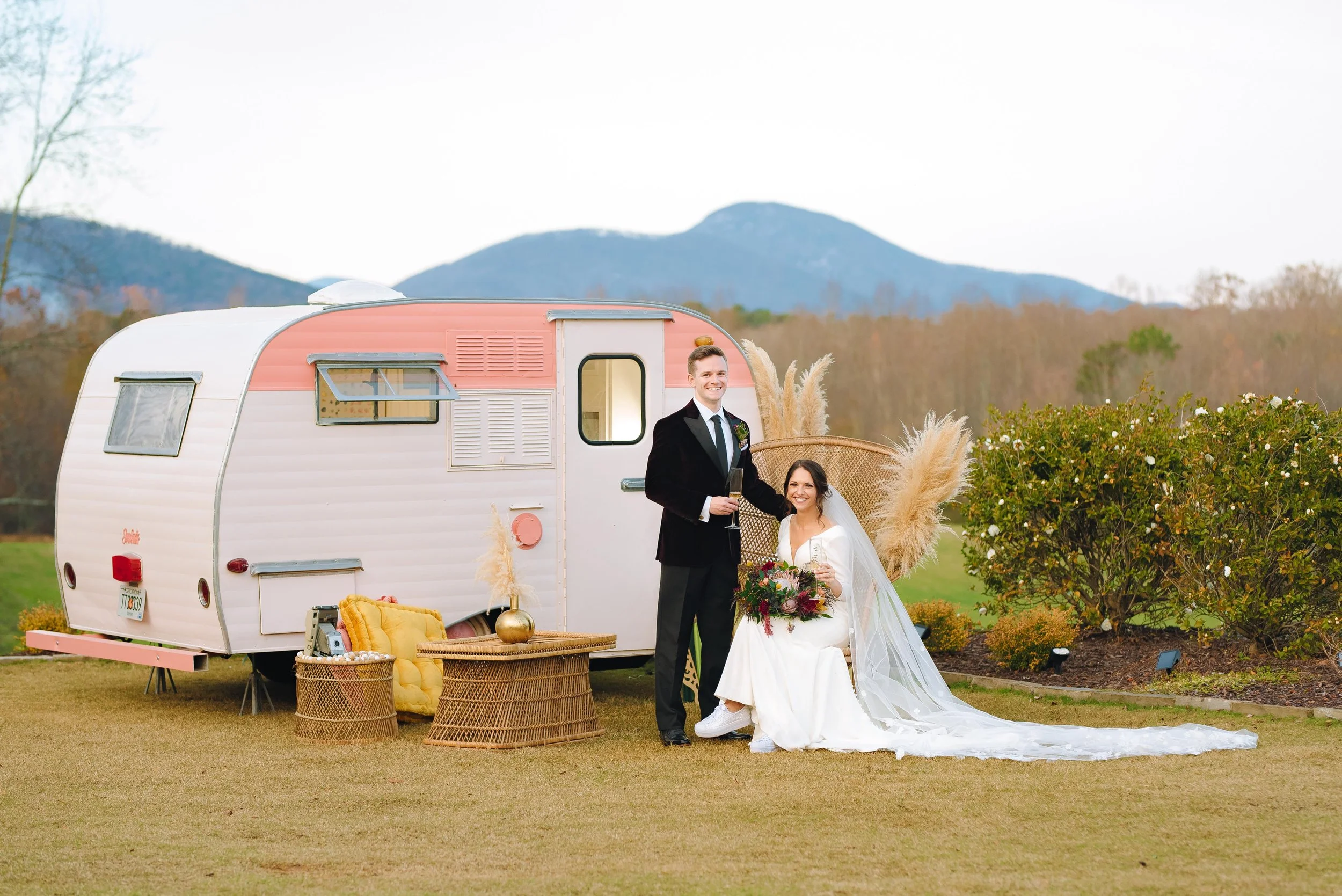 A bride and groom at their wedding outside, standing in front of a vintage camper trailer decorated with natural and bohemian decor, with mountains in the background.  North Georgia Wedding.  Wedding Photography by Sowing Clover Photography.
