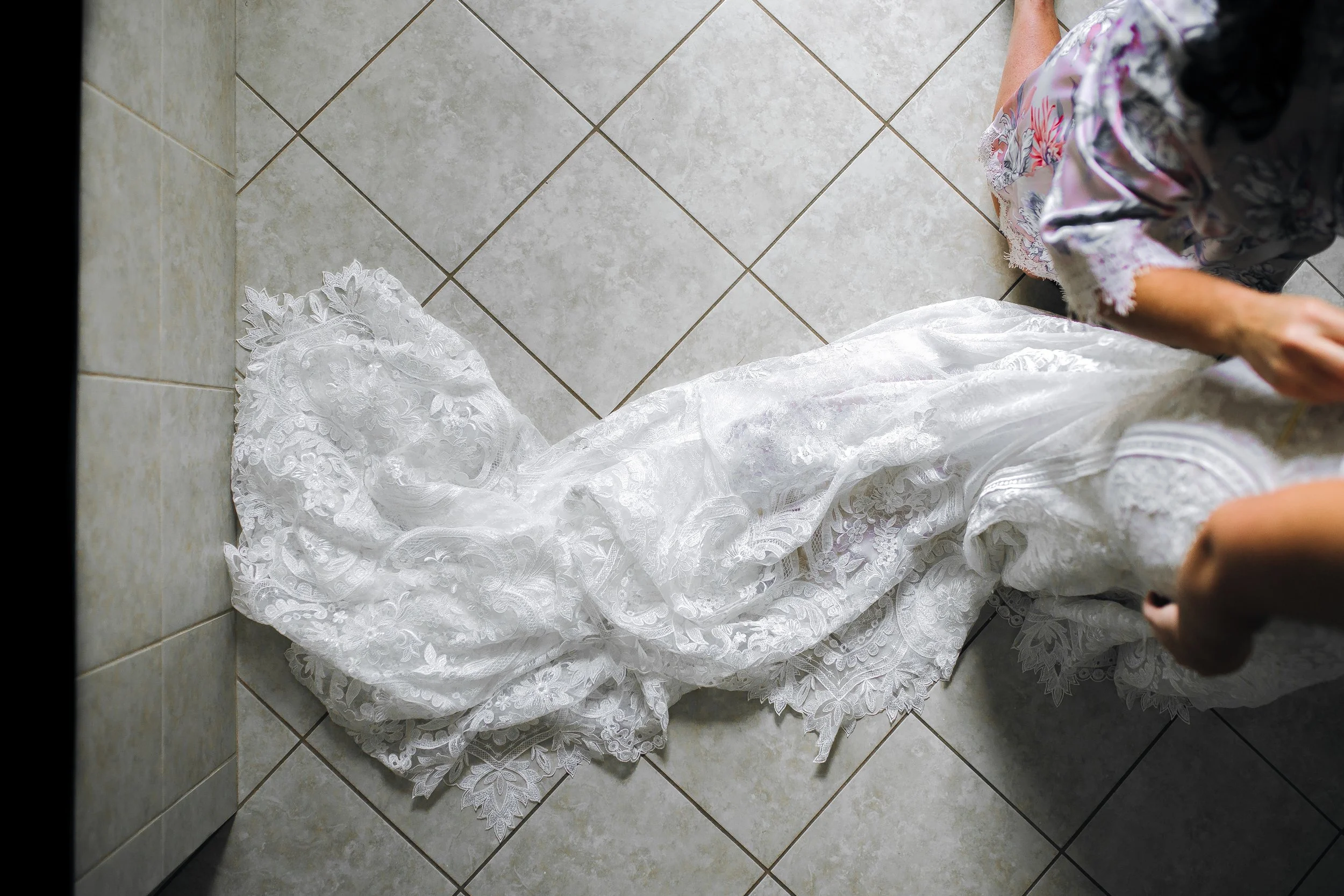 A white lace wedding dress lying on a tiled floor next to a woman in floral pajamas. Wedding Photography by Sowing Clover Photography.