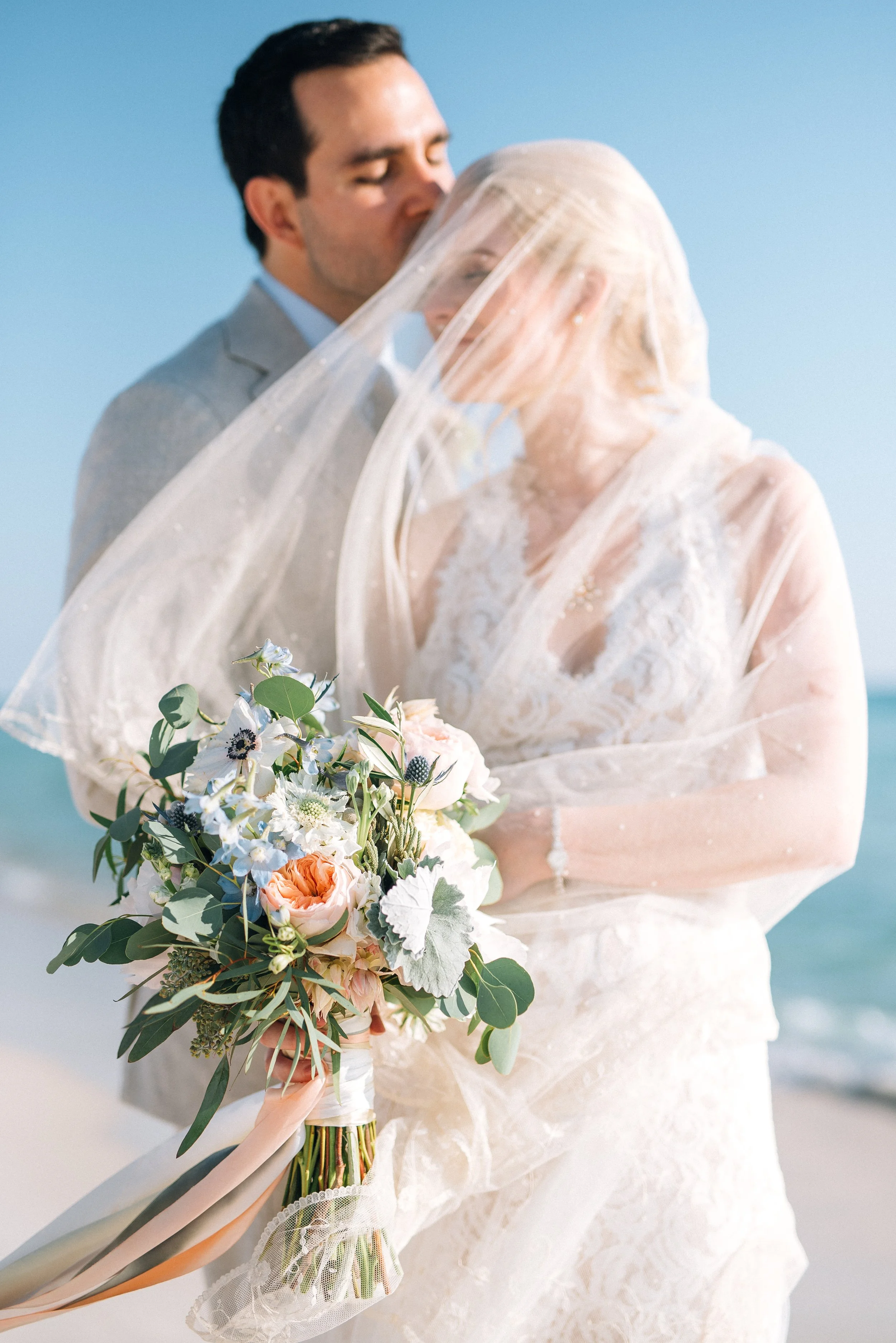 A newlywed couple on the beach with the groom kissing the bride's forehead, the bride holding a bouquet of flowers, under a lace veil. It was a  beach wedding in Florida. Wedding Photography by Sowing Clover Photography.