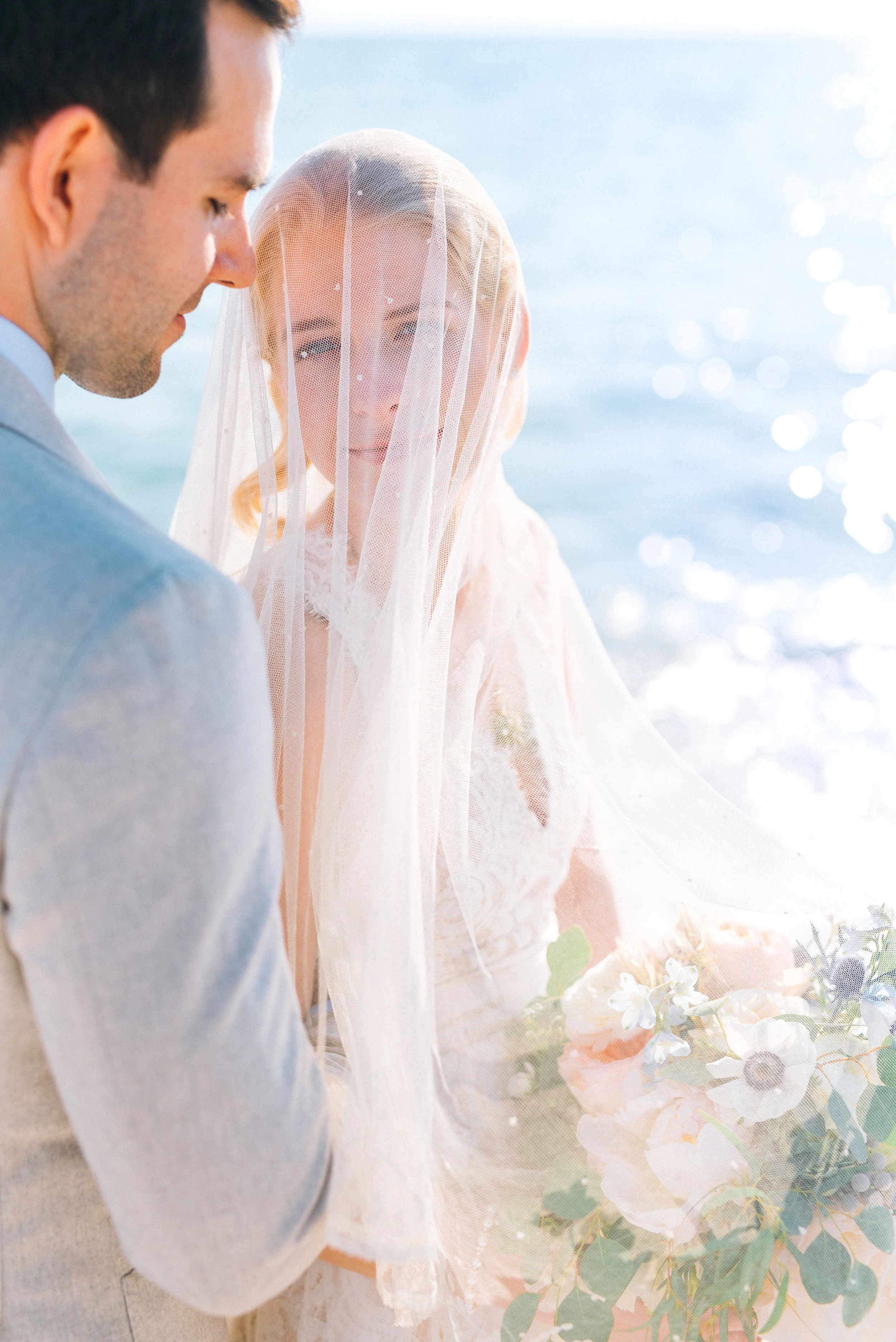 A bride and groom standing outdoors near water, with the bride wearing a veil and holding a bouquet of flowers, and the groom in a suit, both looking at each other. Florida beach wedding.  Wedding Photography by Sowing Clover Photography.