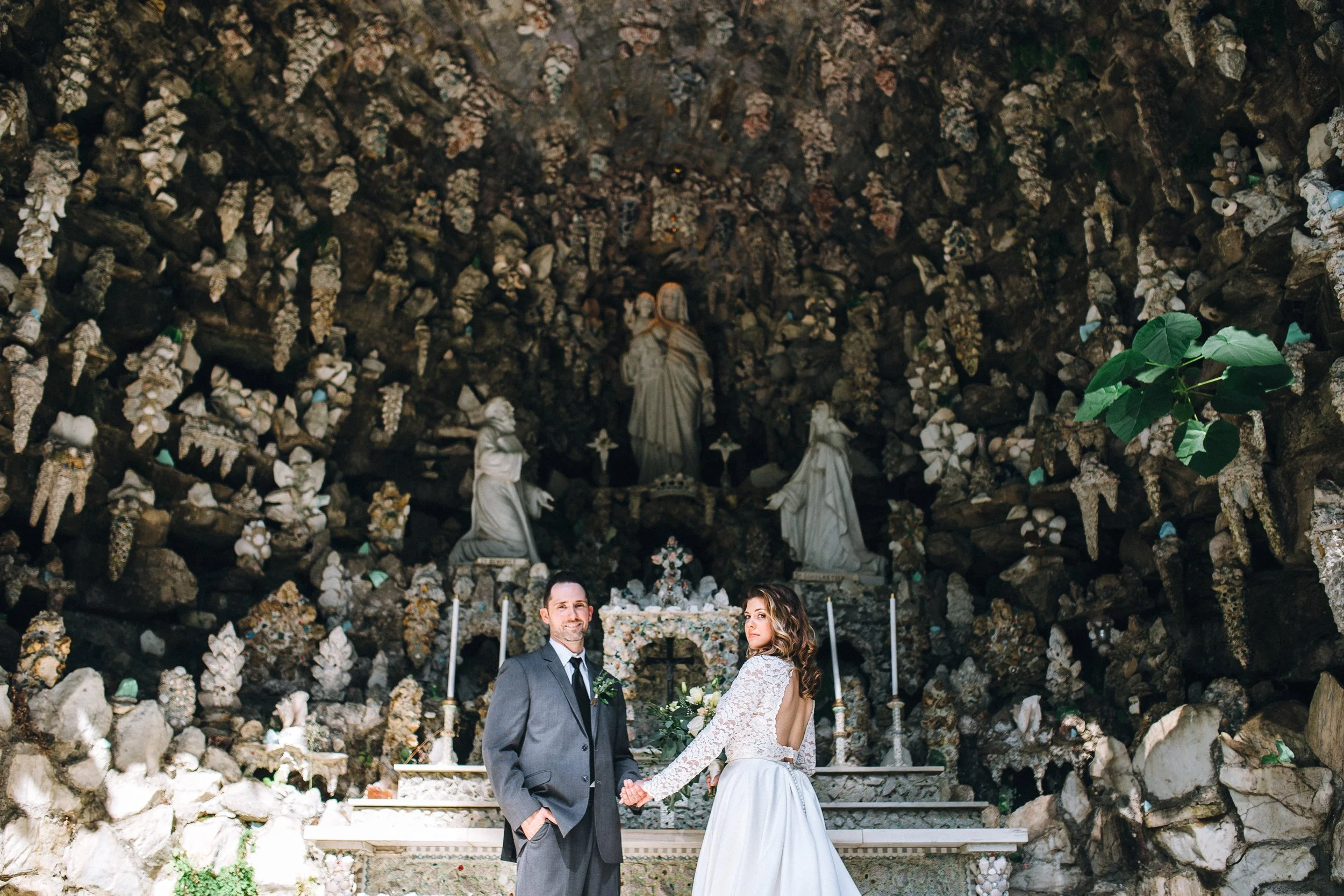 A bride and groom holding hands in front of an altar inside a grotto with religious statues and flowers in Cullman Alabama. Wedding Photography by Sowing Clover Photography.