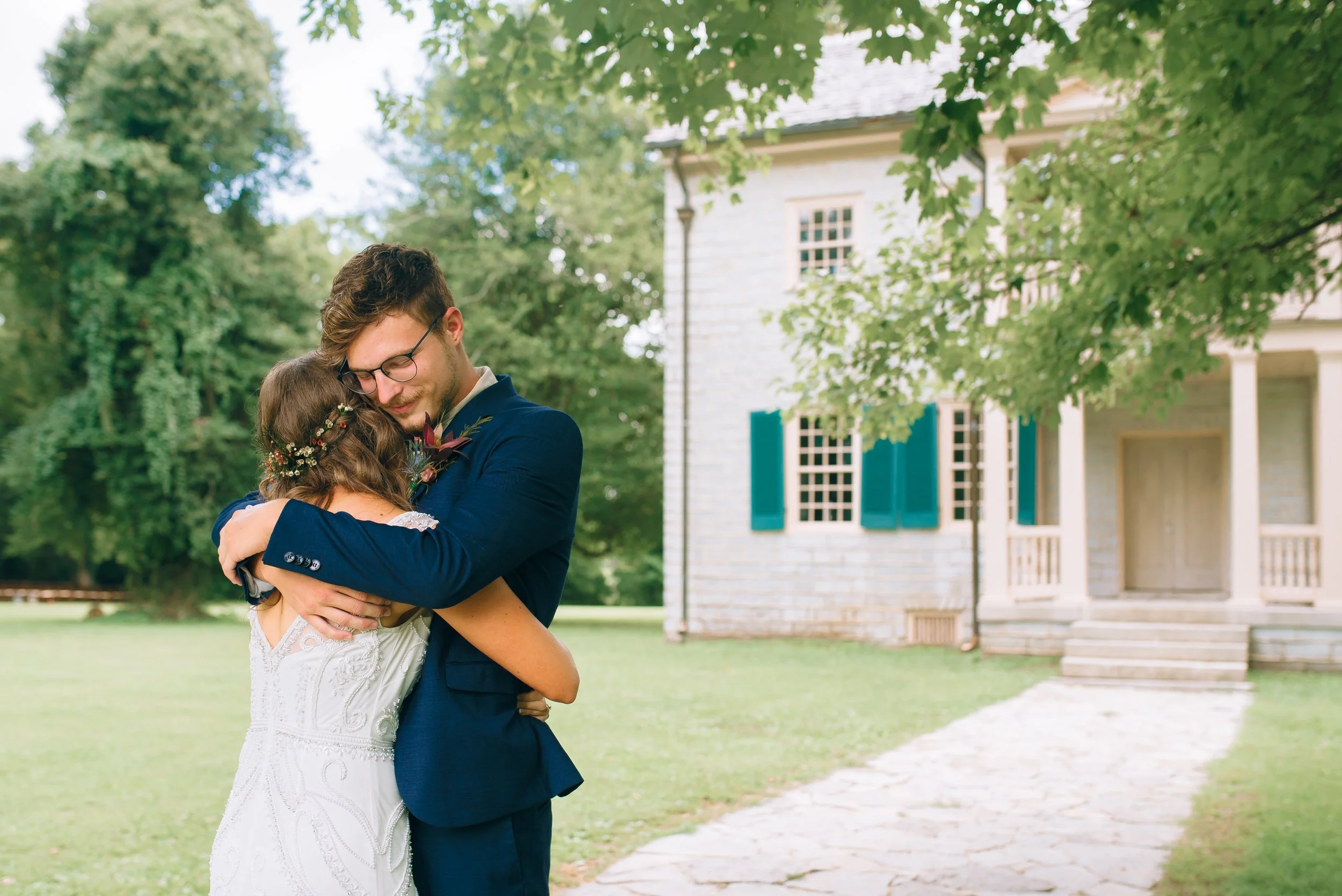 A bride and groom hugging outdoors in front of a vintage house surrounded by trees at Historic Rock Castle in Hendersonville. Wedding Photography by Sowing Clover Photography.
