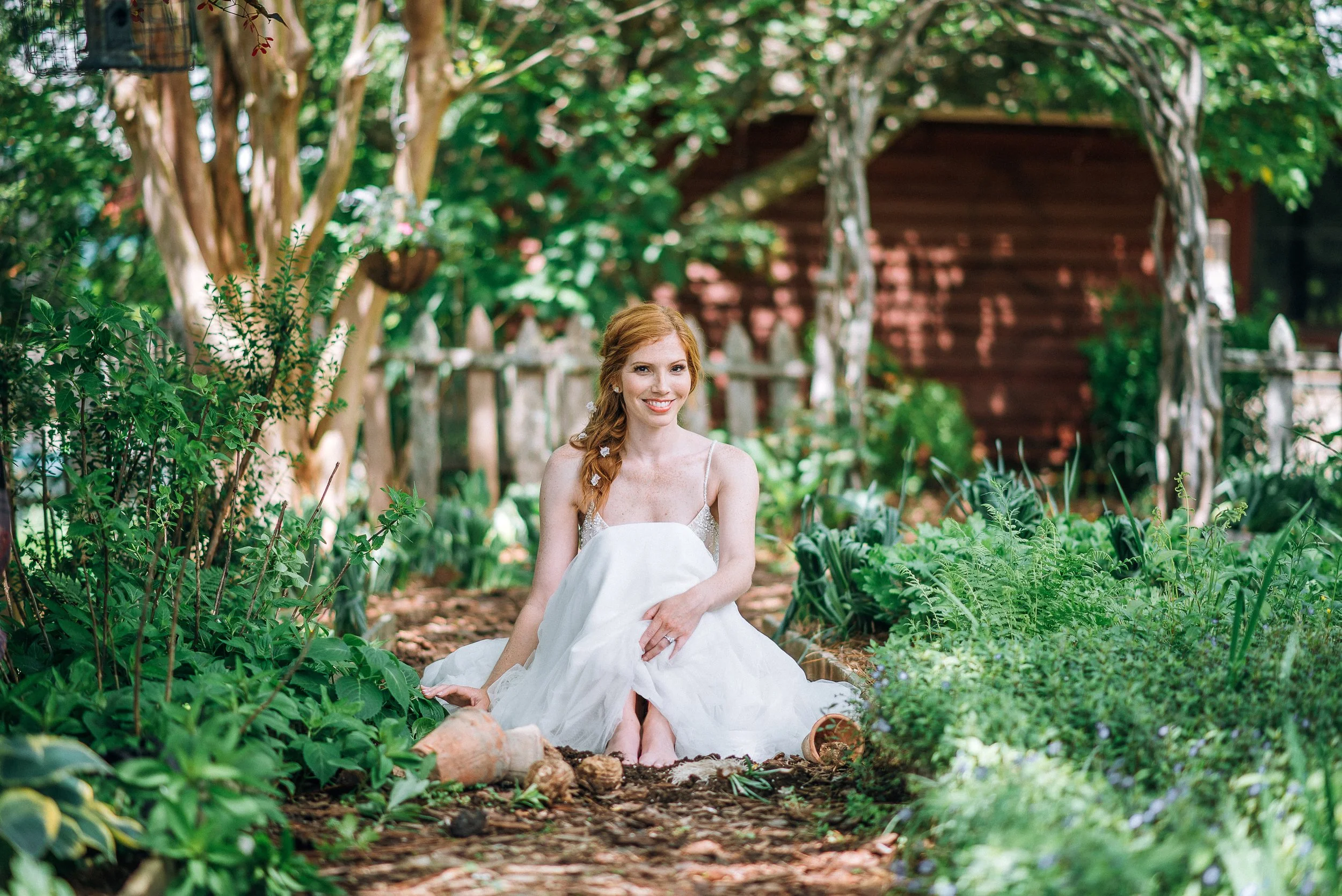 A bride in a white dress sitting on the ground in a lush garden with green plants and trees, a wooden fence, and a red wooden shed in the background at Glen Ella Springs.  Wedding Photography by Sowing Clover Photography..