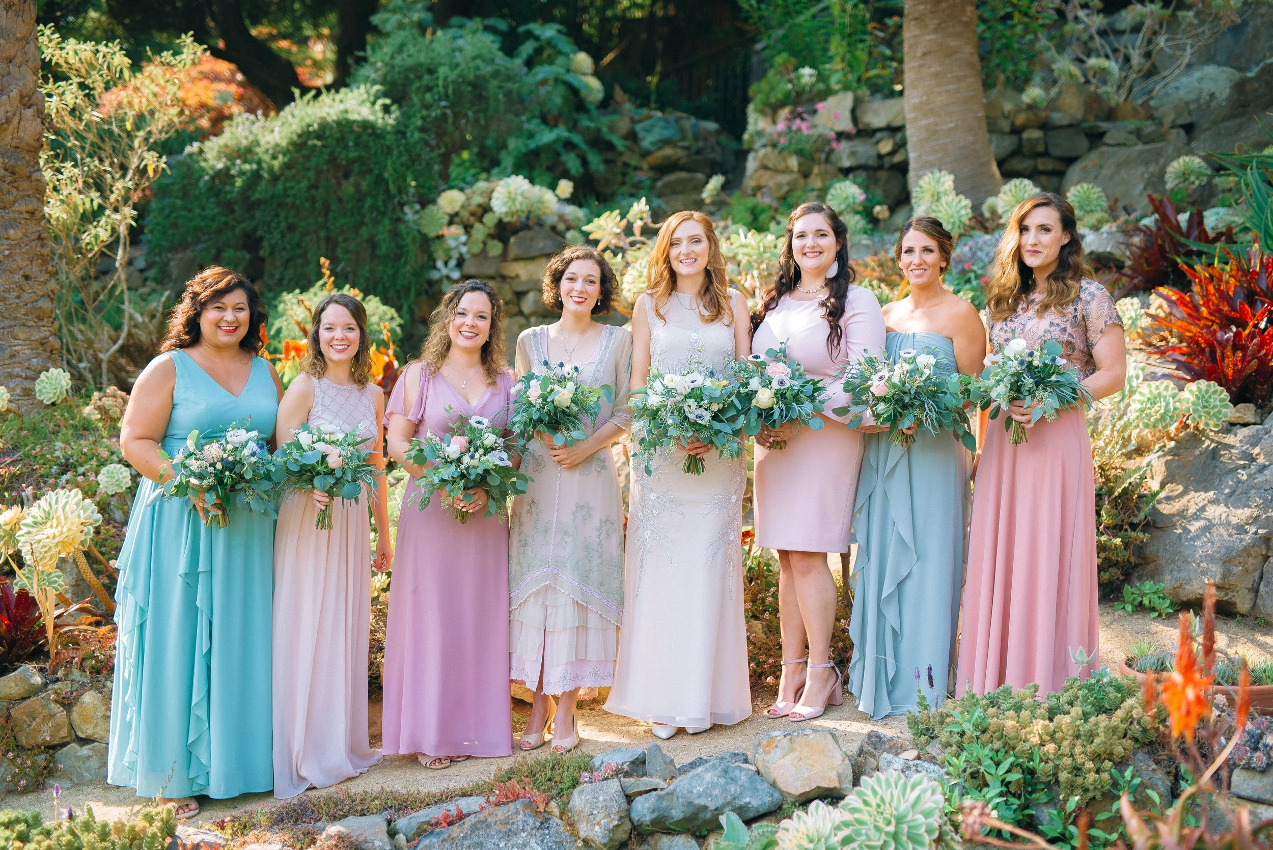A group of eight bridesmaid dressed in pastel-colored dresses, holding bouquets of flowers, standing outdoors in front of a garden with various plants and rocks in San Fransisco. Wedding Photography by Sowing Clover Photography..