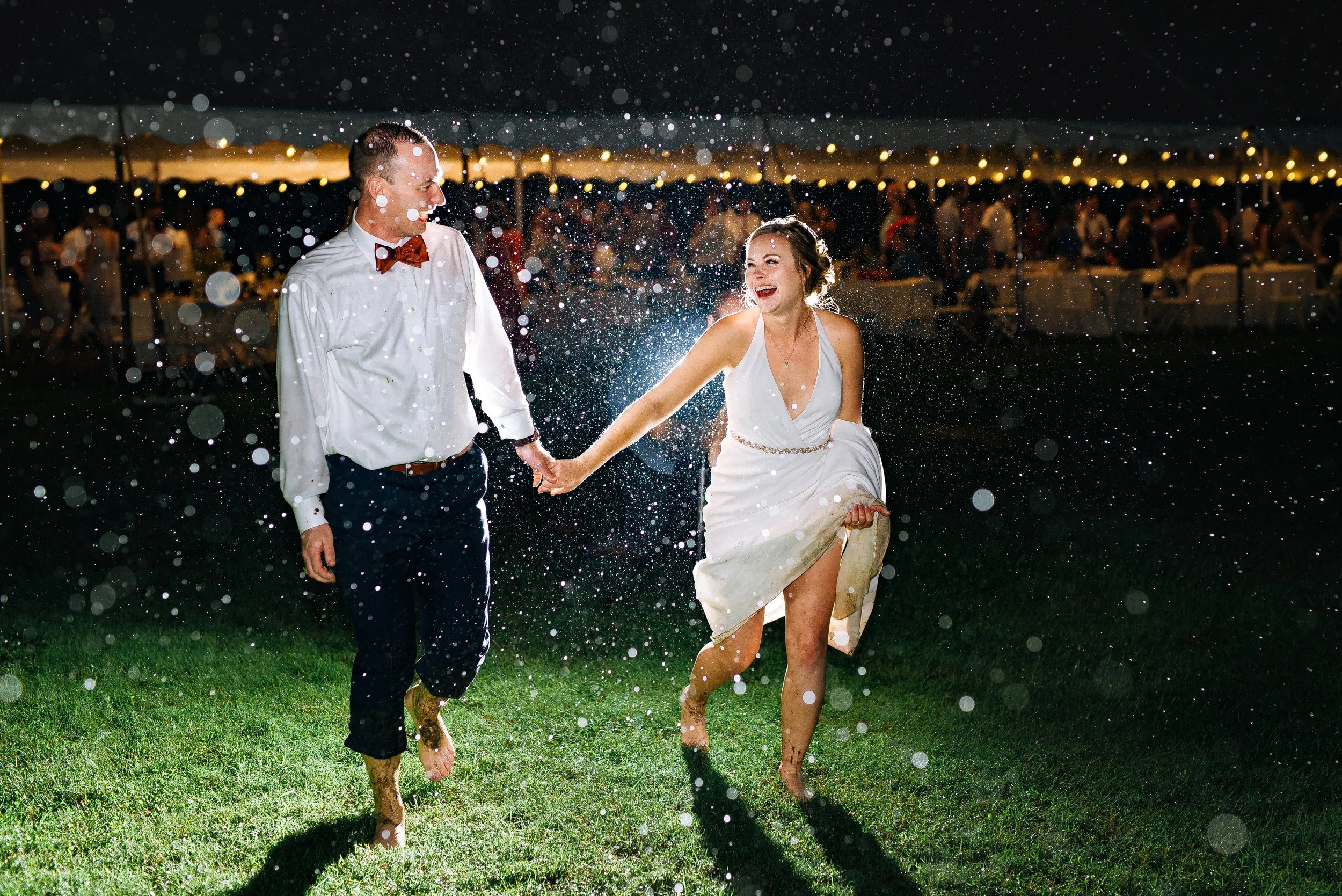 A bride and groom holding hands and laughing while dancing barefoot on grass at night, with water splashing around them and a large tent with string lights in the background. Wedding Photography by Sowing Clover Photography.