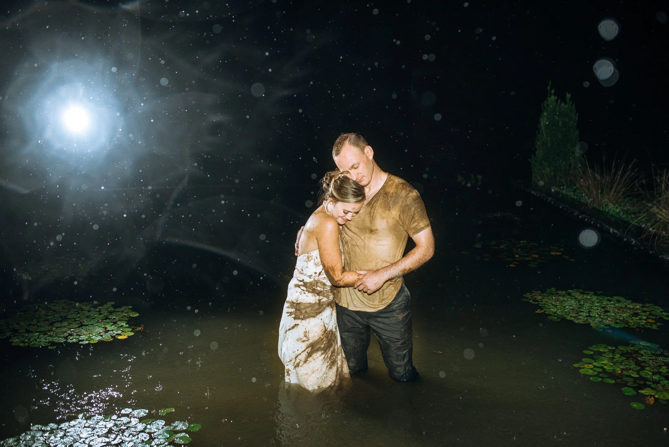 A bride and a groom as a couple standing together in a body of water at night, holding hands and embracing, with the woman leaning her head on the man's shoulder, both appearing muddy and wet in Nashville. Wedding Photography in by Sowing Clover