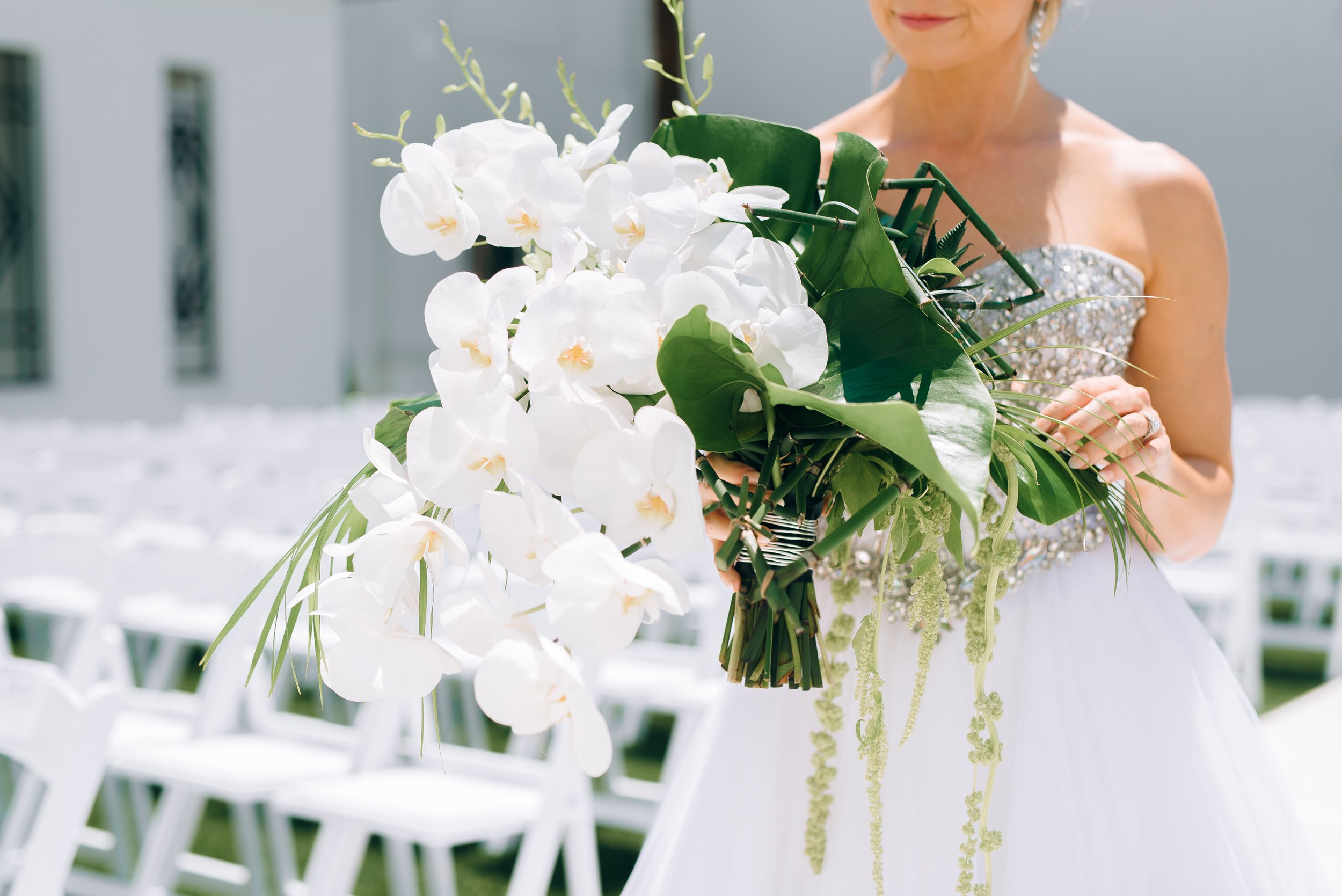 A bride holding a large bouquet of white orchids and green foliage, standing outdoors with white chairs in the background at The Clay Theater in Green Cove Springs, Florida. Wedding Photography by Sowing Clover Photography.