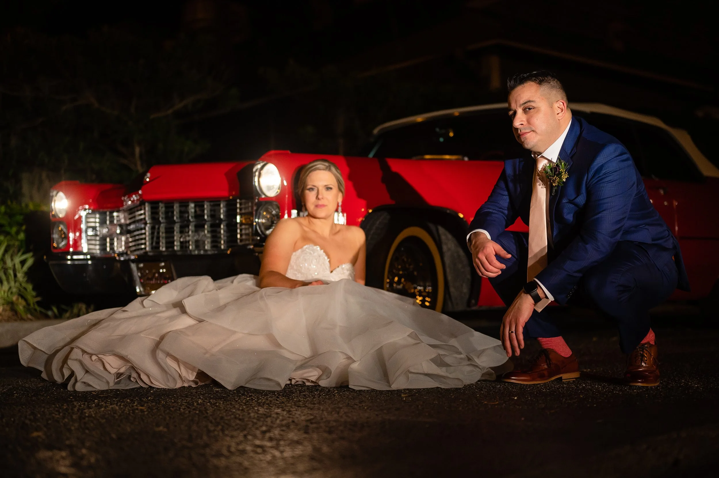 A couple dressed in wedding attire poses in front of a classic red car at night. The woman is sitting on the ground in a white wedding gown, while the man is crouching next to her in Atlanta wedding venue. Wedding photography by Sowing Clover Photo