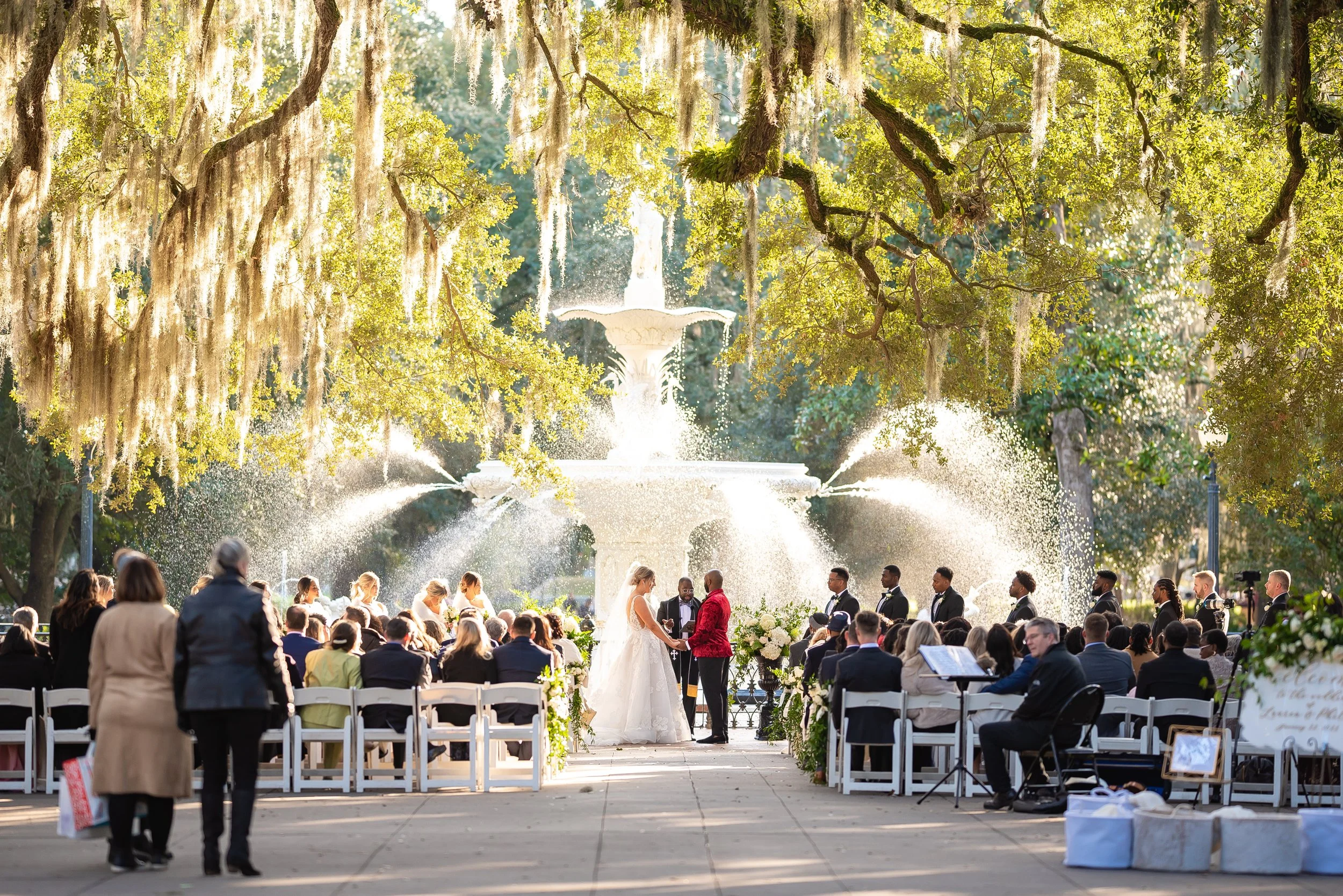 A wedding ceremony taking place outdoors near a fountain in a park, with the bride and groom exchanging vows under large trees with hanging moss in front of the fountain at Forsyth Park in Savannah, Georgia. Wedding Photography by Sowing Clover Photo