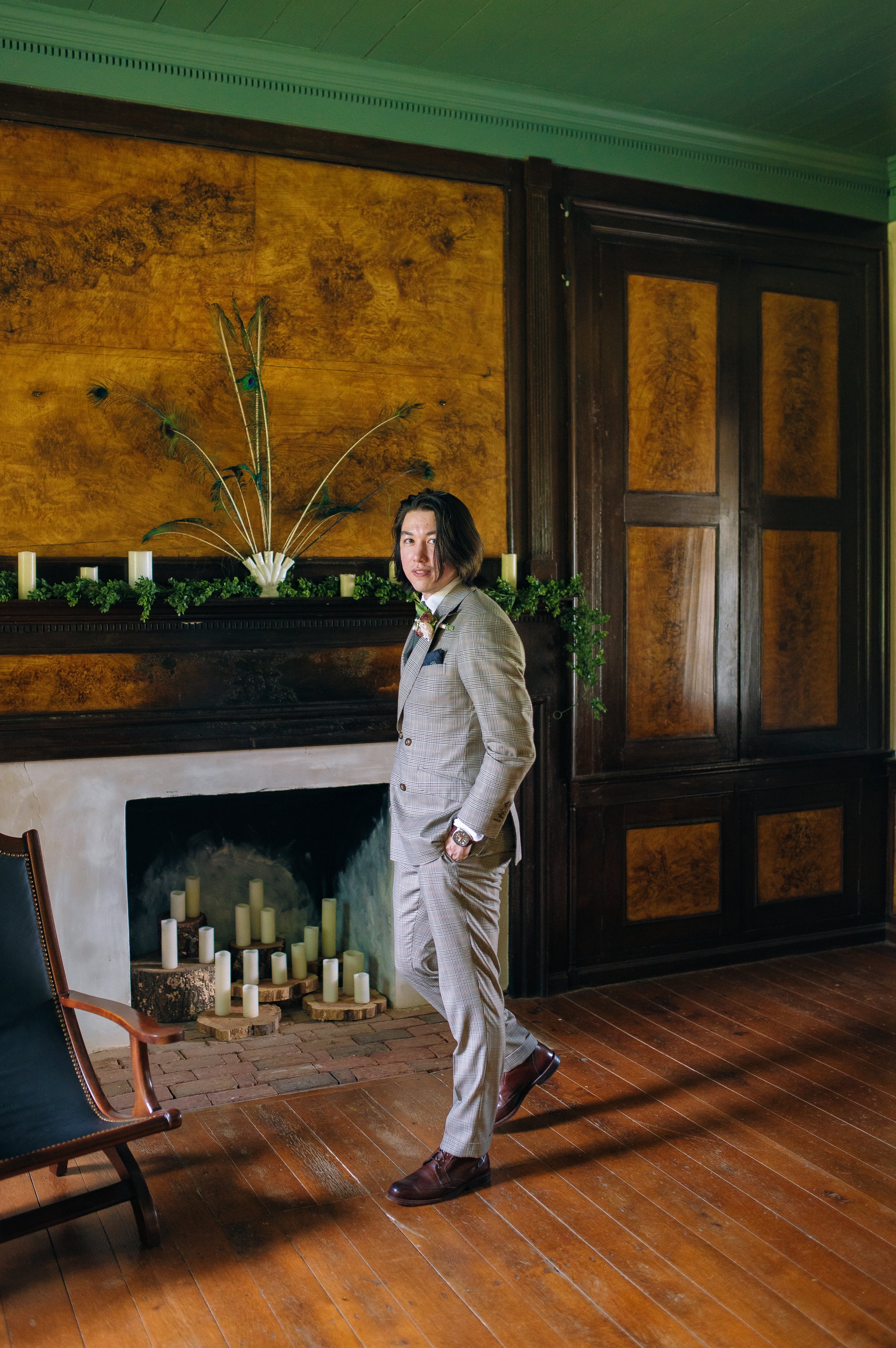 A groom with shoulder-length dark hair dressed in a gray checked suit posing indoors near a fireplace with candles at Rock Castle Wedding Venue in Nashville. Wedding Photography by Sowing Clover Photography.