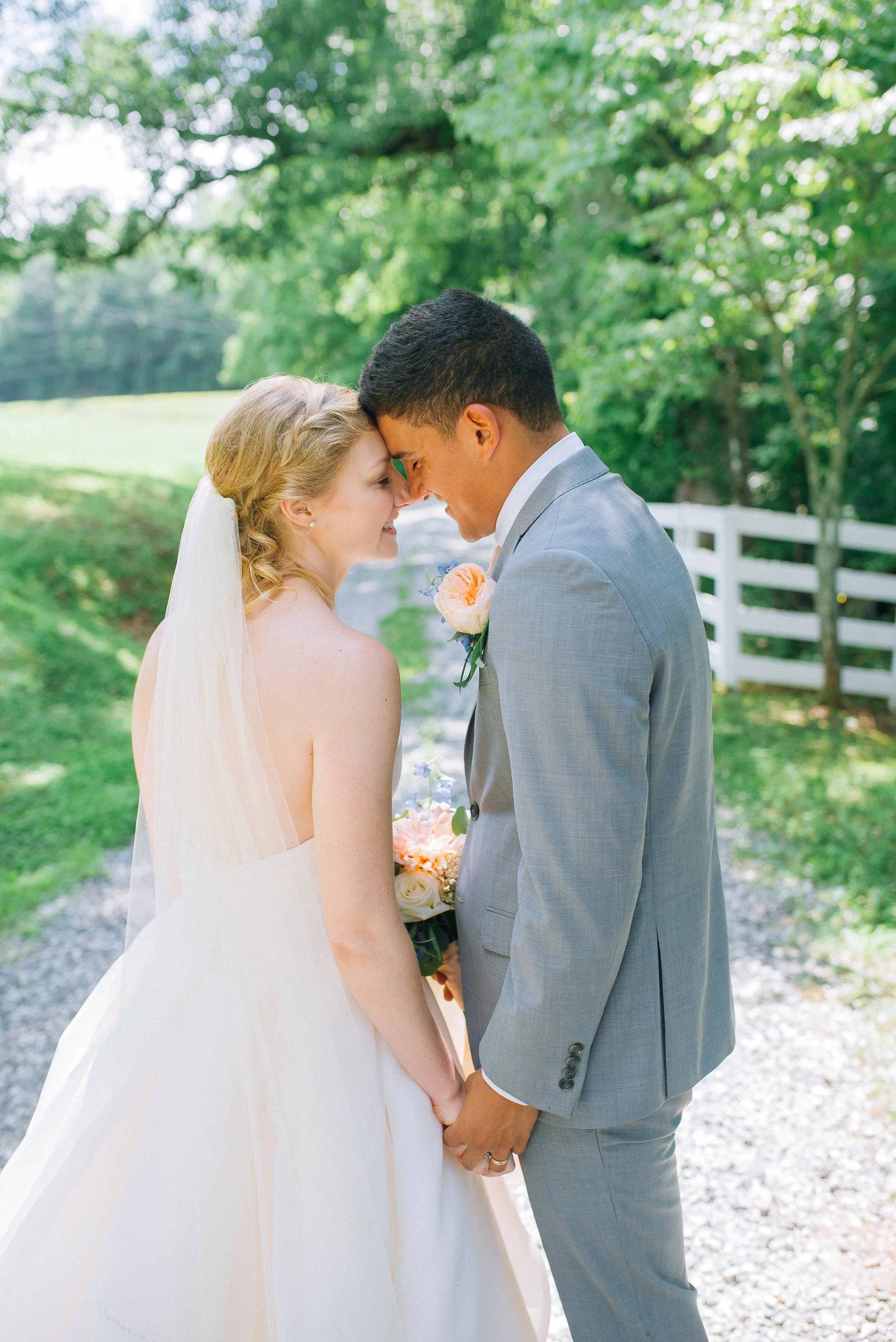 Bride and groom with foreheads touching, holding hands, outdoors with trees, grass, and a white fence, Wedding photography by Sowing Clover Photography.
