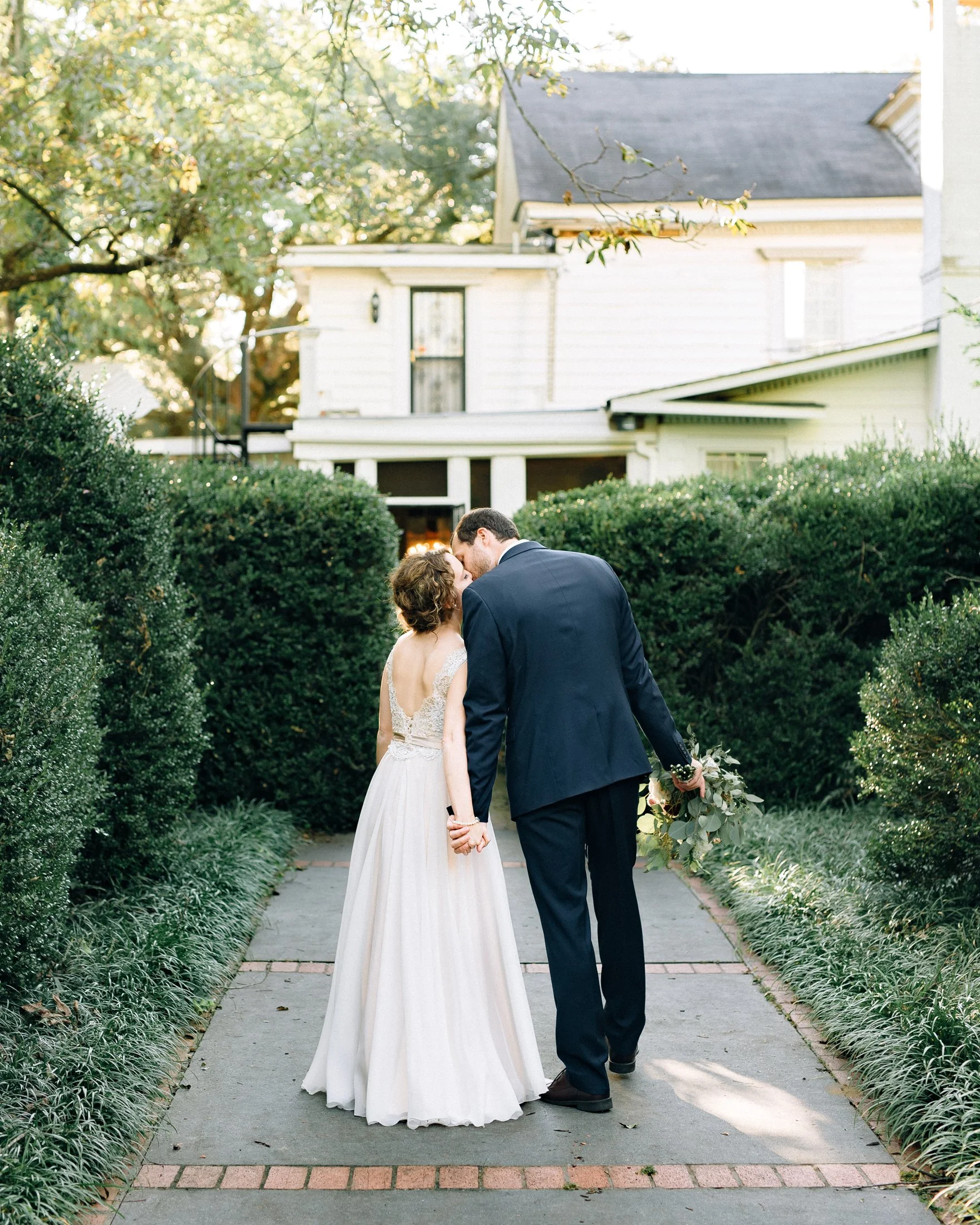 A newlywed couple holding hands and sharing a kiss on a garden pathway at sunset, with a white house and green bushes in the background in Nashville TN. Wedding Photography in by Sowing Clover Photography