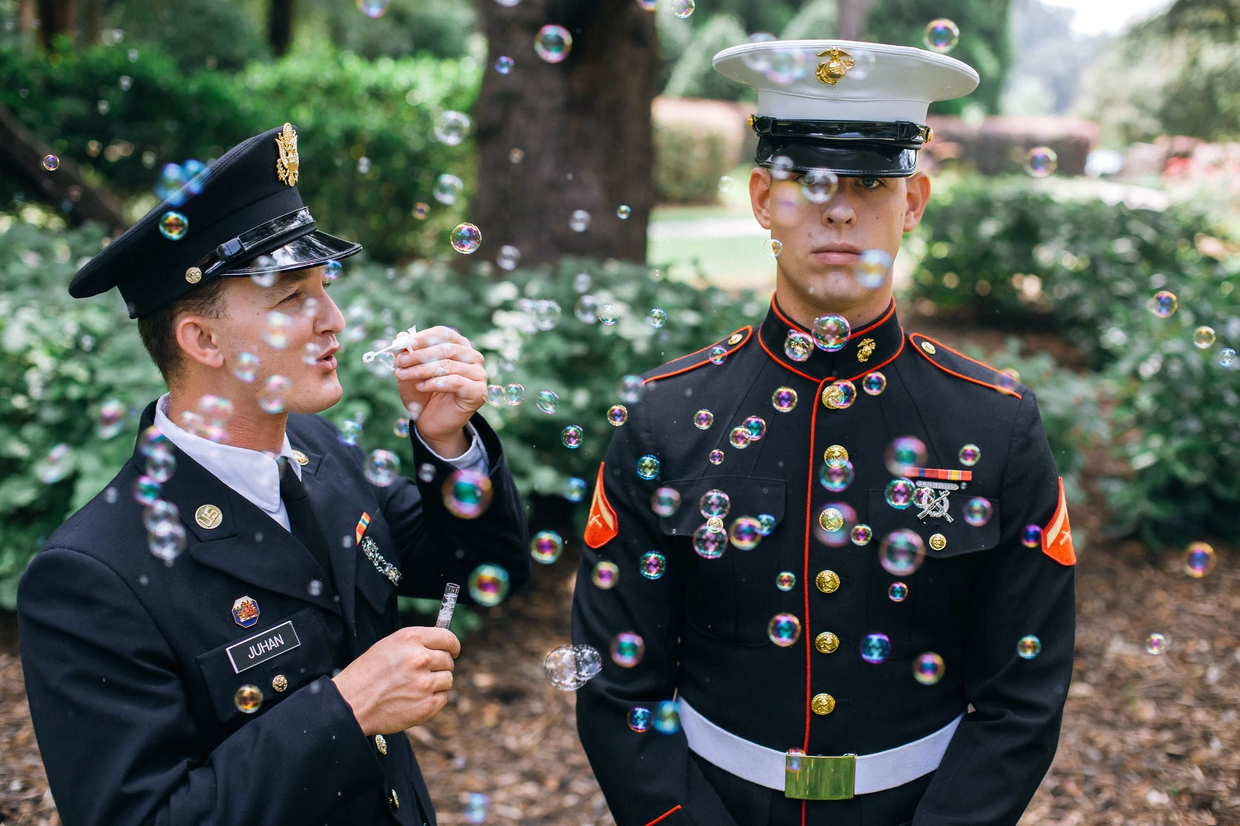 Two U.S. Marines in dress uniforms standing outdoors among trees and greenery, surrounded by floating soap bubbles. One of them is a groom in Atlanta Georgia. Wedding photography by Sowing Clover Photography.