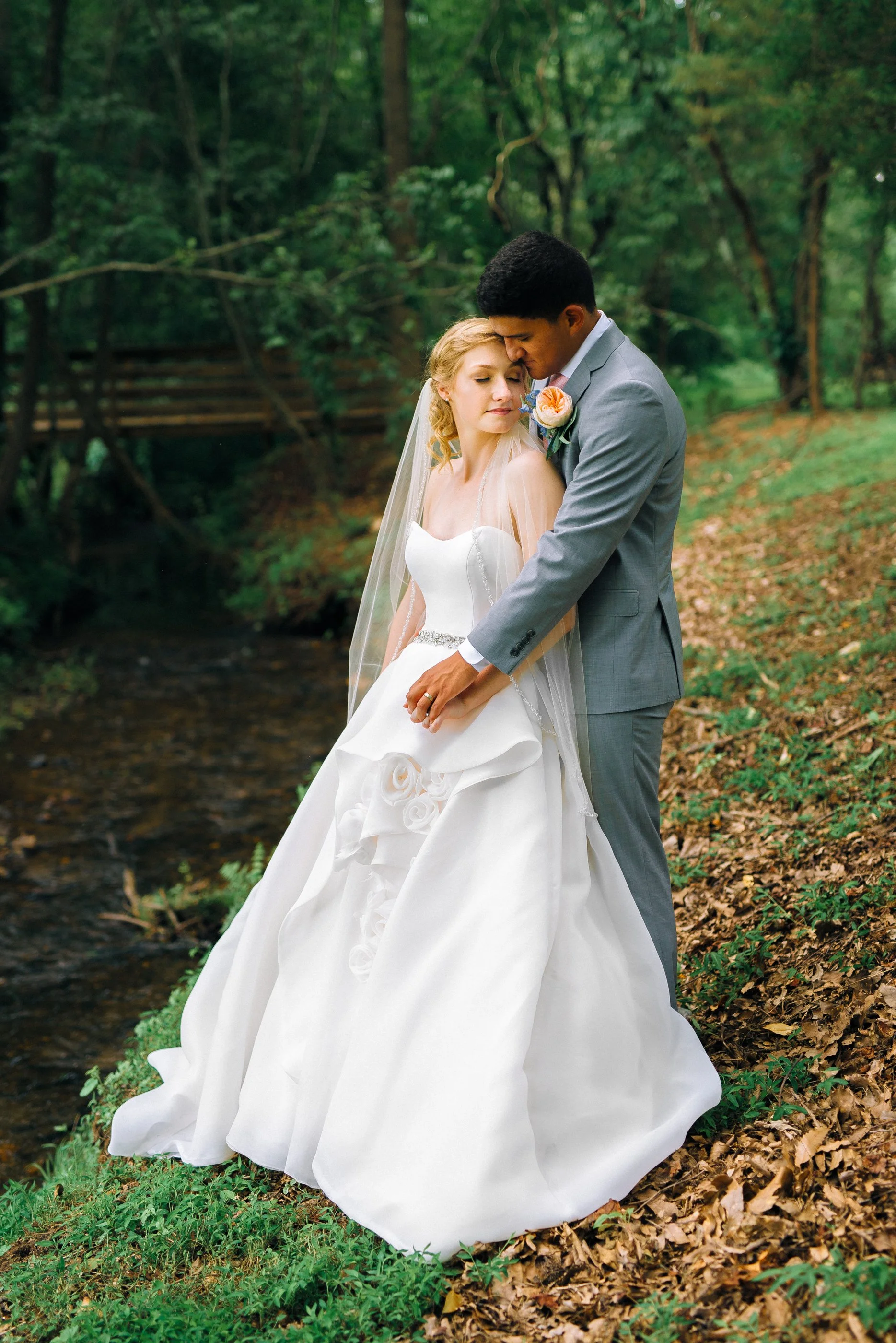A bride and groom stand close together in a wooded outdoor setting, with the groom gently holding the bride's hand and resting his forehead on hers. Memphis TN, Wedding photography by Sowing Clover Photography.