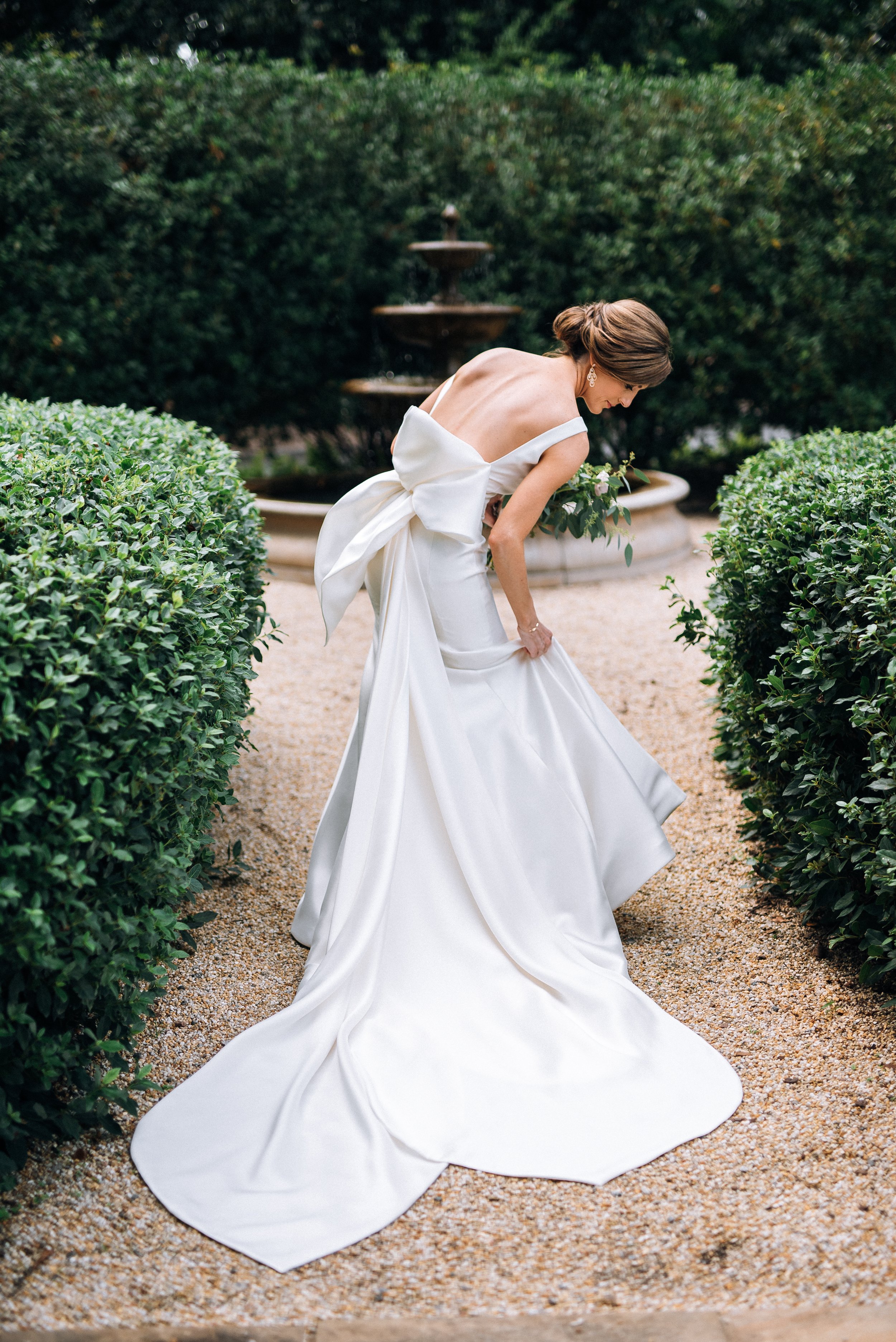 Bride in a white wedding gown with a large bow at the back, standing outdoors near a fountain, holding a bouquet of greenery, with a hedge and fountain in the background at The Estate in Atlanta. Wedding photography by Sowing Clover Photography
