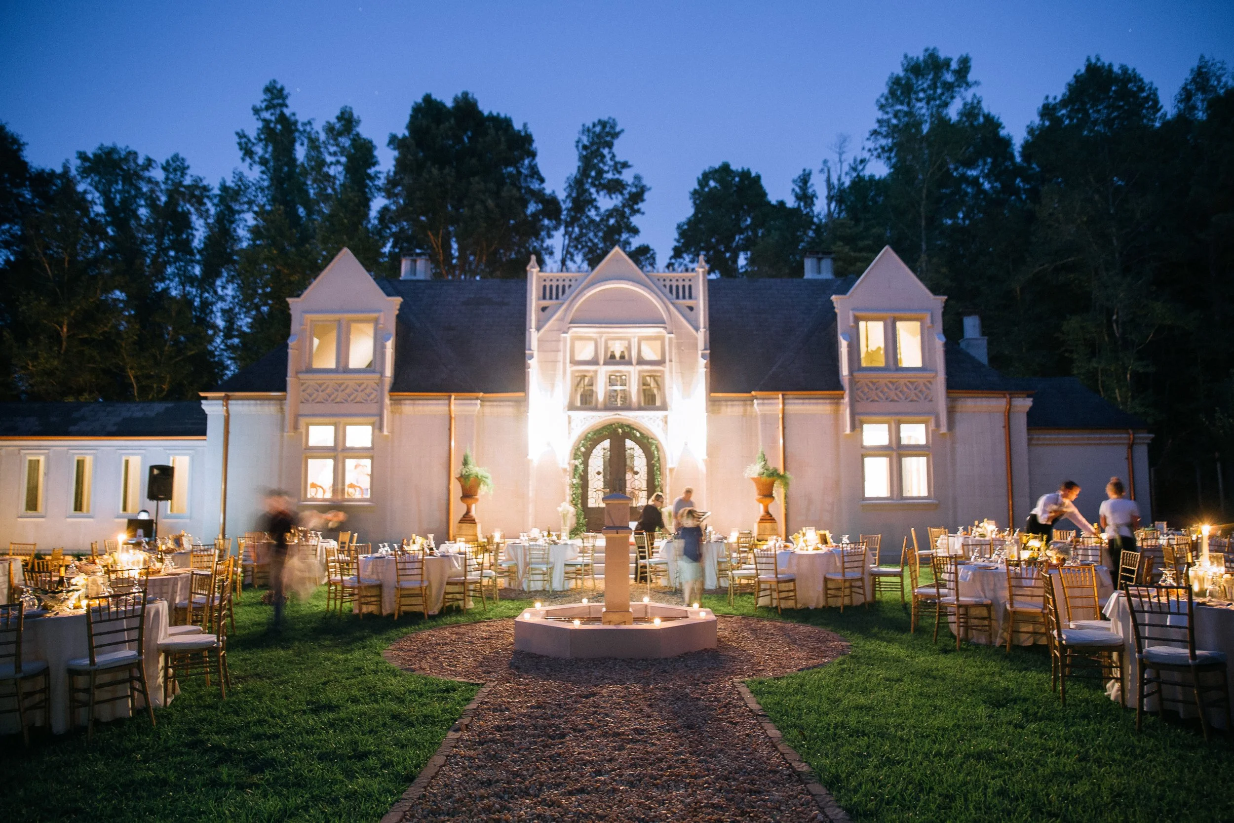 An outdoor wedding reception setup at night with tables and chairs arranged on a lawn in front of a large mansion, illuminated by string lights and candles Atlanta Georgia Wedding photography by Sowing Clover Photography.