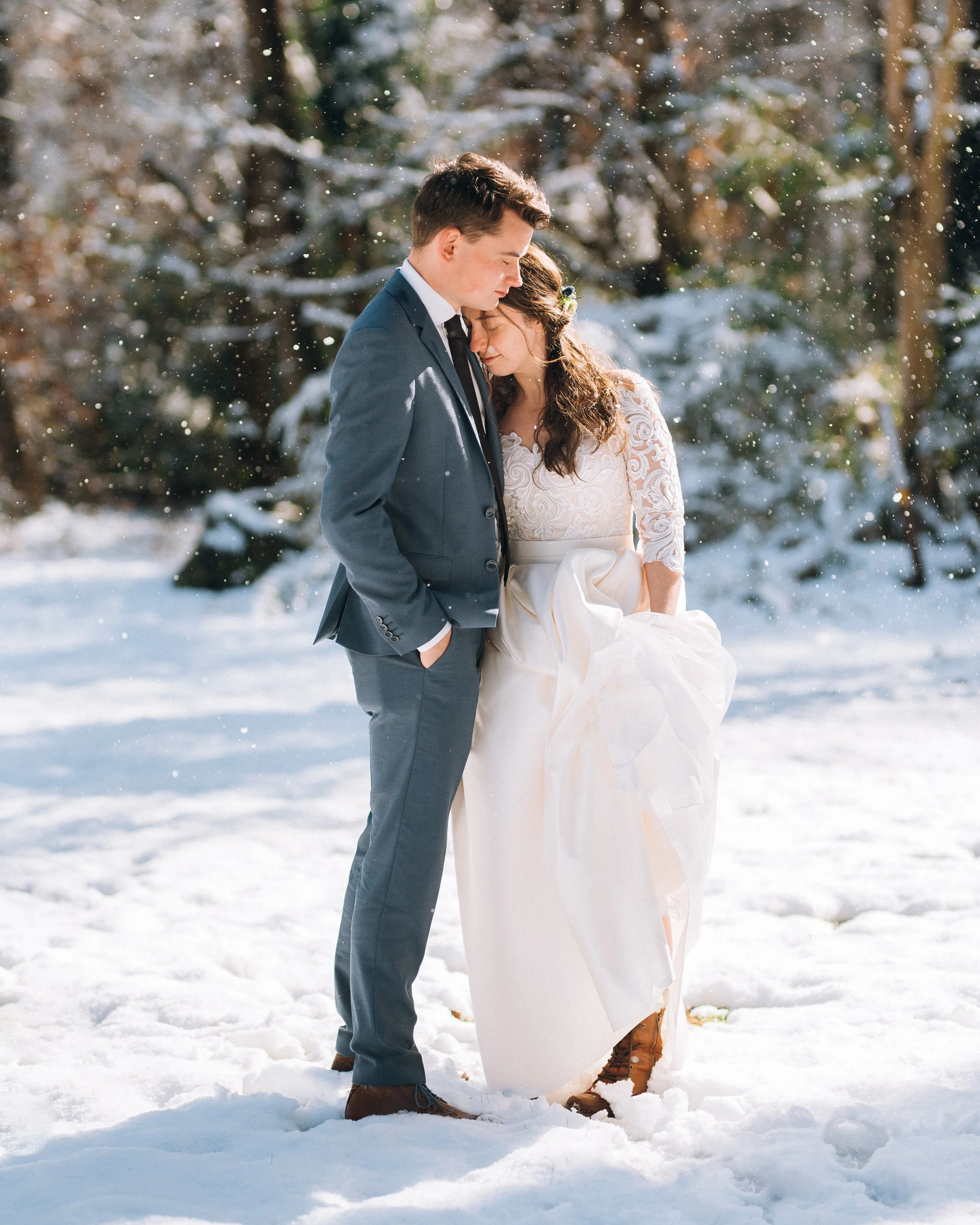 A couple dressed in wedding attire standing closely together in the snow with a snowy forest background. Snowy Atlanta December Wedding. Wedding photography by Sowing Clover Photography.