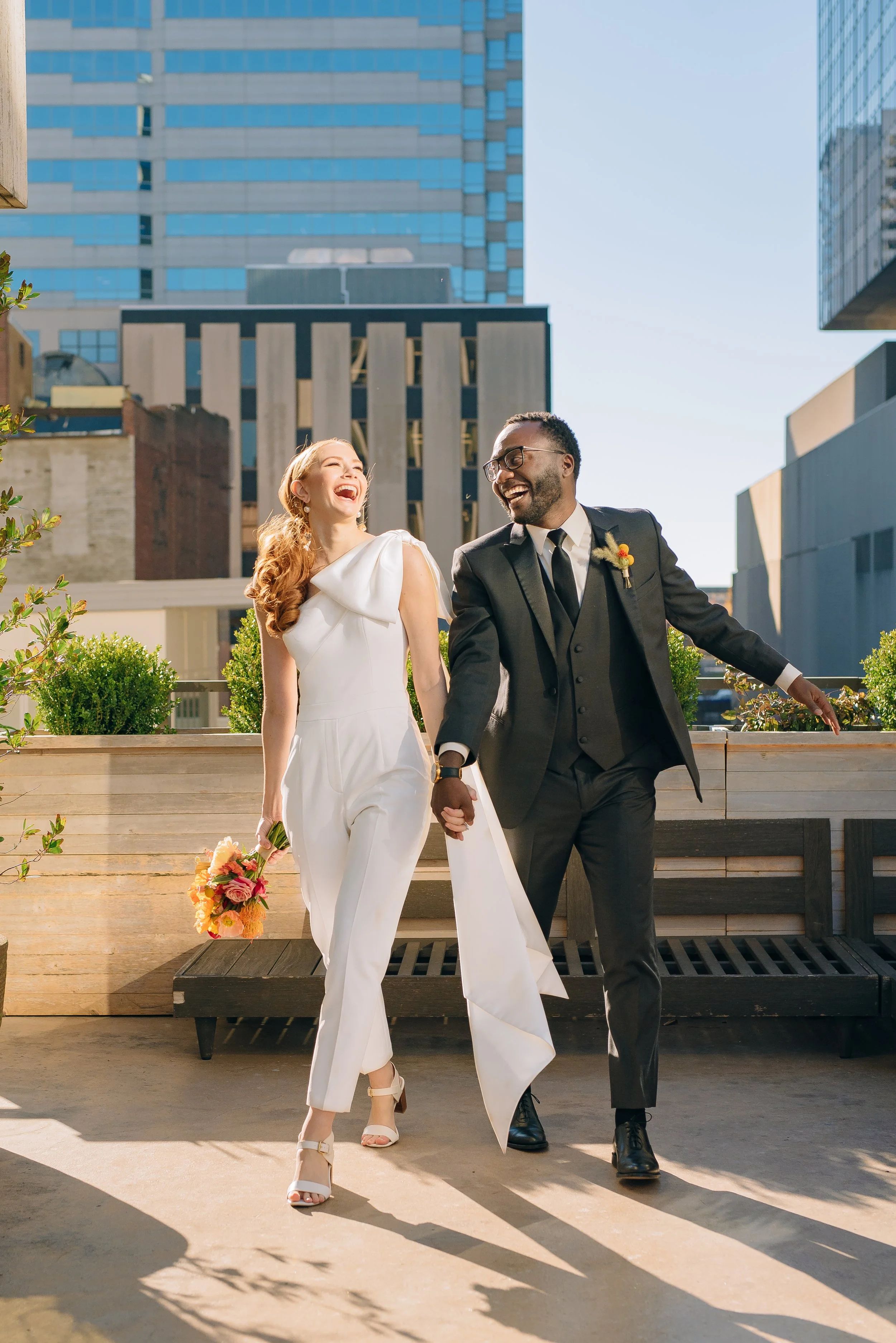 A happy couple, a bride in a white dress holding a bouquet and a man in a black suit, is walking hand in hand on a rooftop in an urban setting, smiling and laughing at the Fairlane Hotel. Wedding Photography by Sowing Clover Photography.