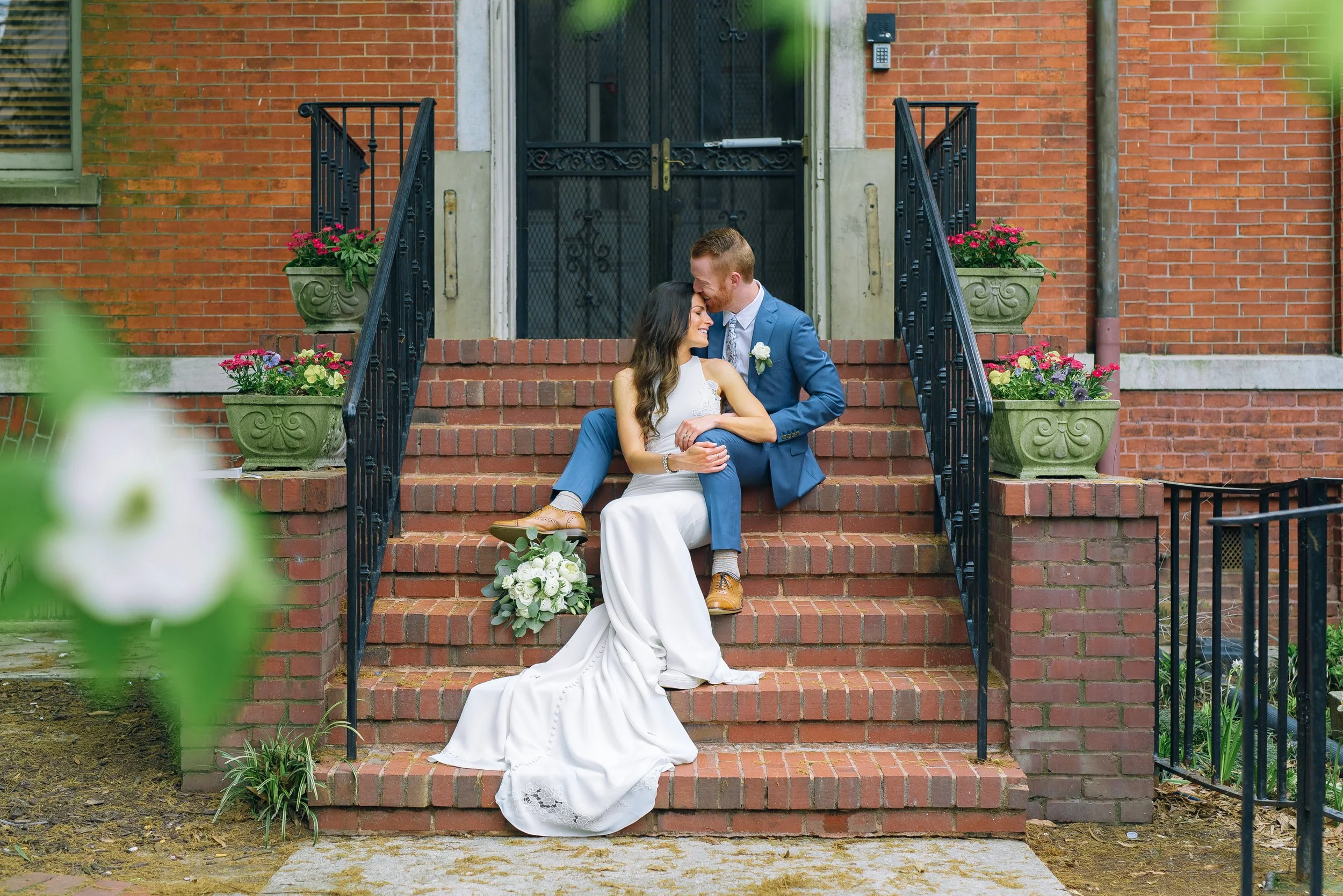 A bride and groom sitting on brick stairs outside a building, sharing a romantic moment. The bride wears a white dress with a train, holding a bouquet of white flowers. The groom wears a blue suit with a boutonniere. Colorful potted flowers line the 