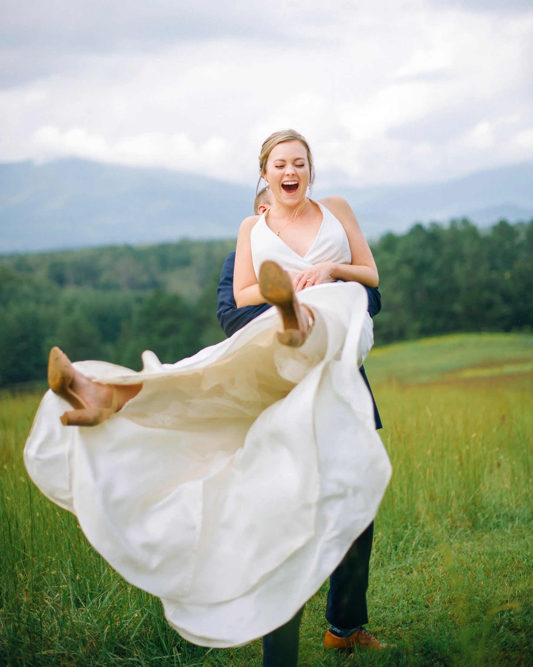 A bride in a wedding dress being lifted and held by a man in a suit in a grassy field with mountains and cloudy sky in the background. Wedding Photography by Sowing Clover Photography.