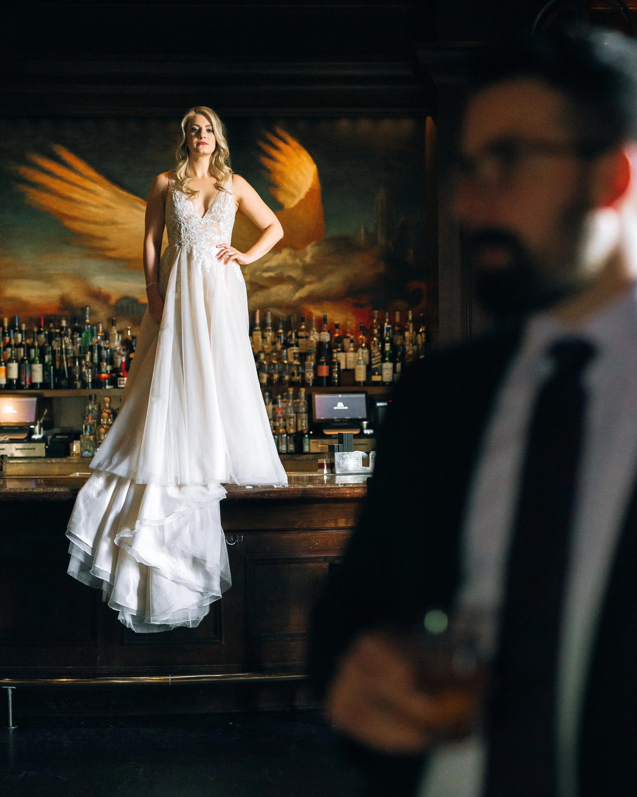 A woman in a white wedding dress stands on a bar counter, with a large bottle cap with wings painted on the wall behind her. In the foreground, a groom with a suit at the Ritz in Atlanta. Wedding Photography by Sowing Clover Photography.
