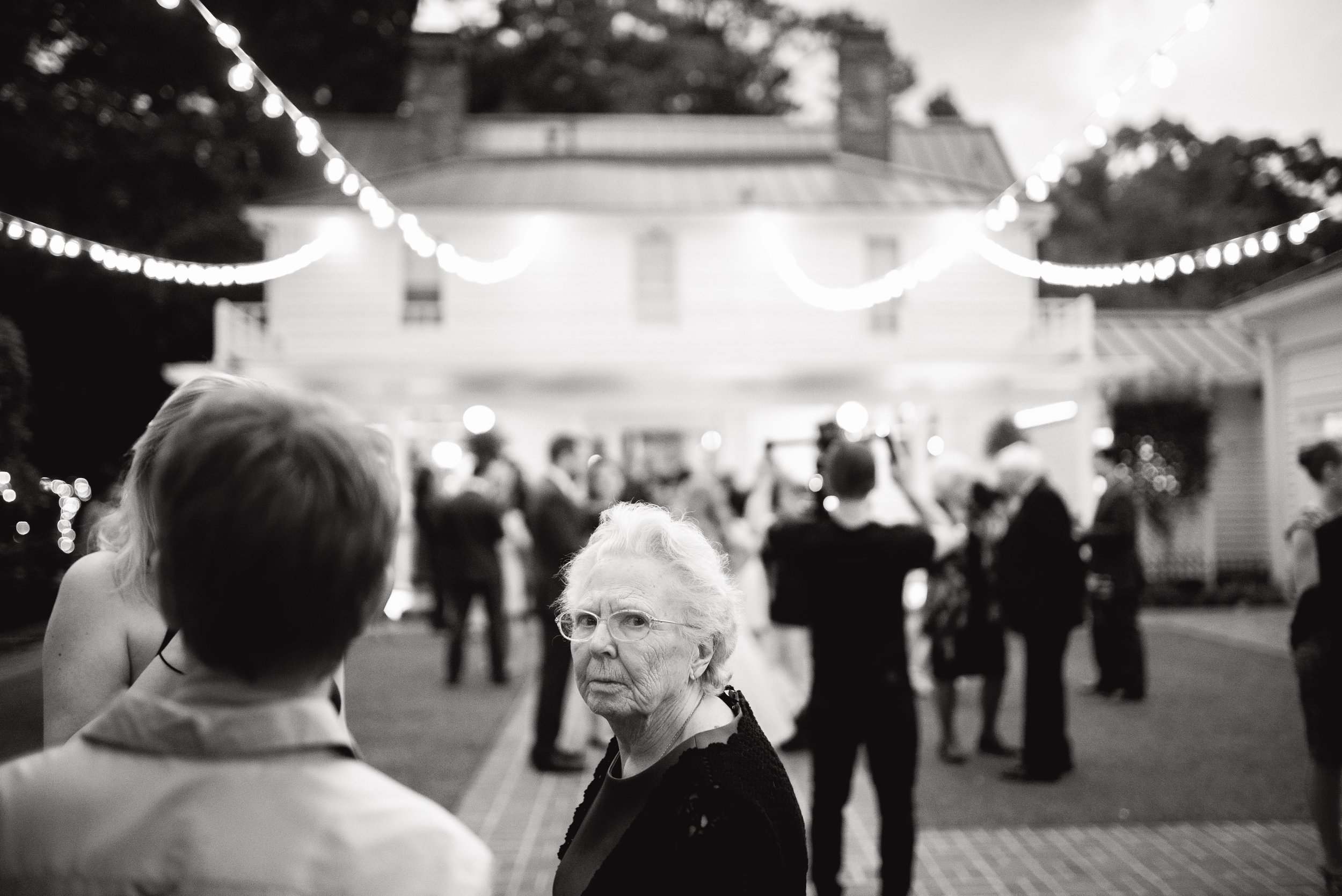 An elderly woman with glasses looks at the camera during a weather reception outdoors at night, with string lights and a house in the background, and several people mingling. Atlanta. Wedding Photography by Sowing Clover Photography.