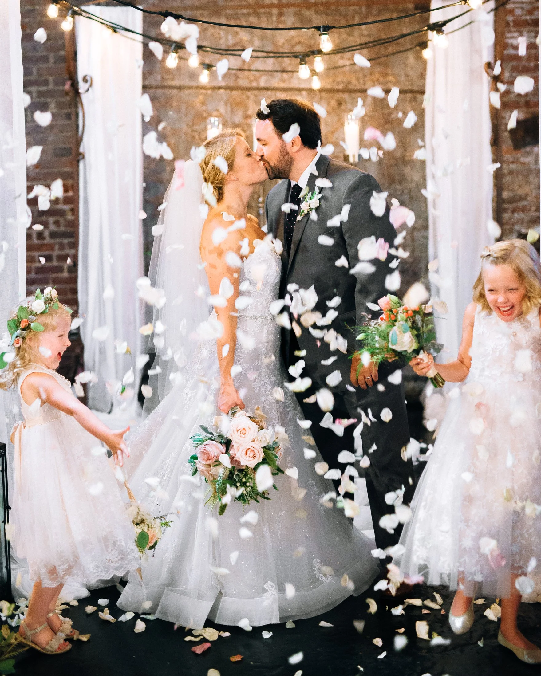 A newlywed couple kisses under falling flower petals while two young girls, dressed in white, celebrate with bouquets during a wedding reception in an industrial-style venue at King Plow. Wedding Photography by Sowing Clover Photography.