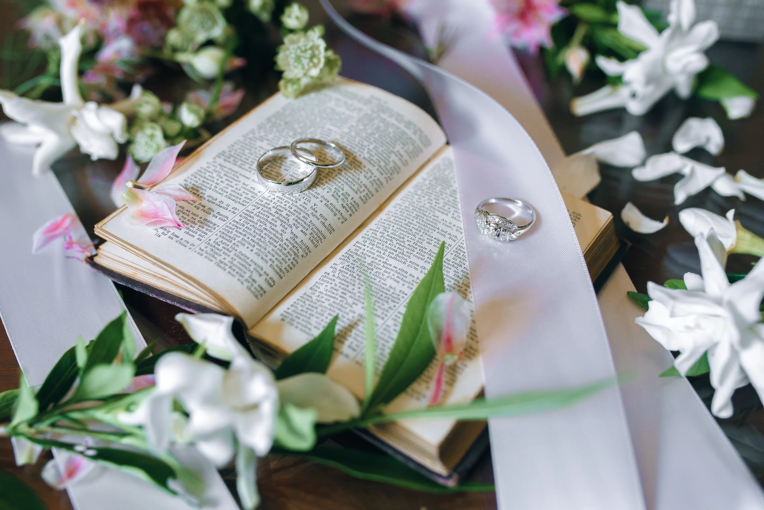 Open Bible with wedding rings on the pages, surrounded by white and pink flowers and greenery, with white ribbon decoration in Atlanta Georgia Wedding photography by Sowing Clover Photography