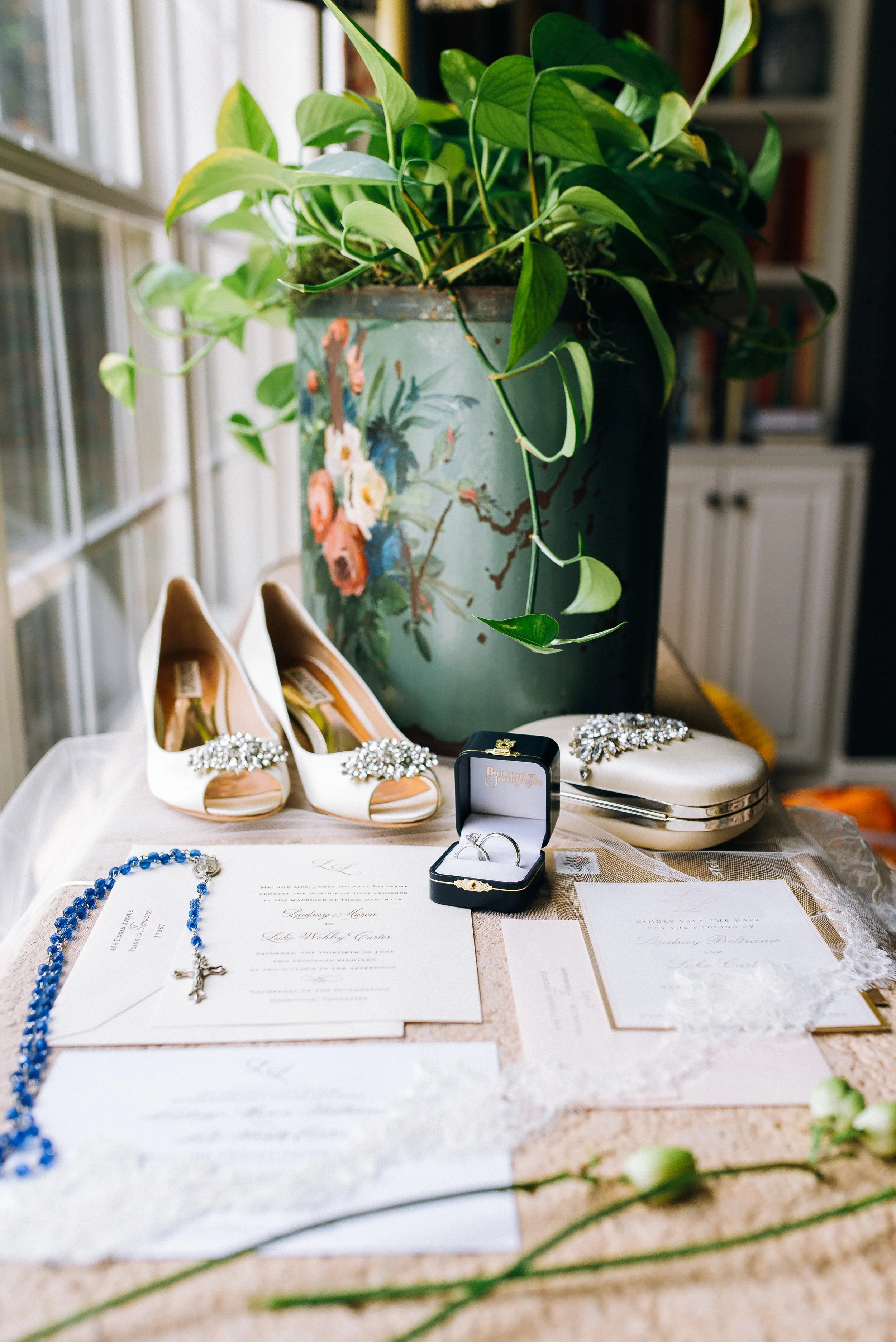 Wedding invitation, engagement ring, pearl clutch, blue rosary with silver cross, white bridal shoes with rhinestone embellishments, floral decor and greenery in a vase on a table in Nashville. Wedding Photography in by Sowing Clover Photography